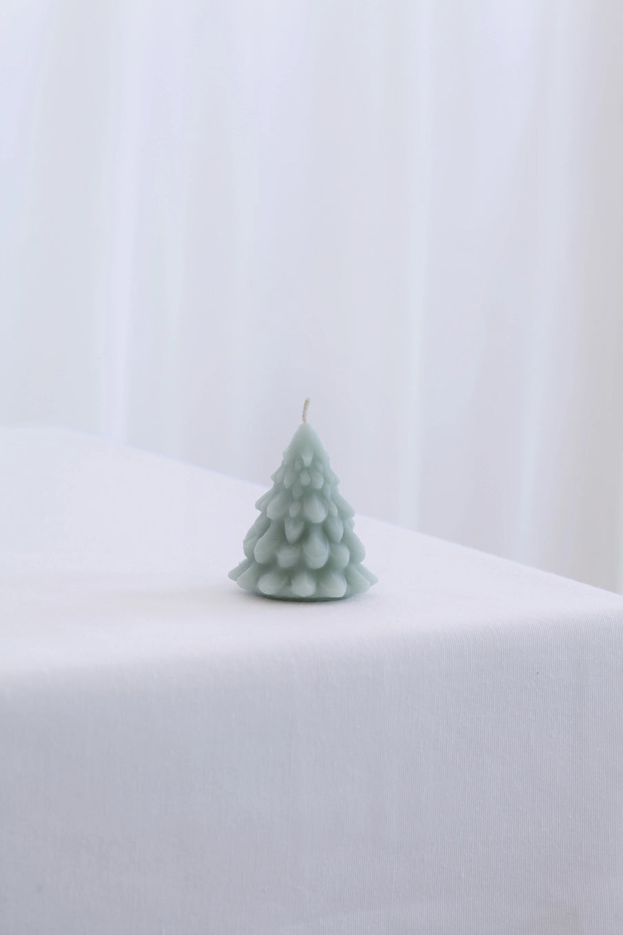 A small tree-shaped candle in a pale sage green color, standing on a white draped tablecloth against a soft white curtain background. ©Mo&Co.