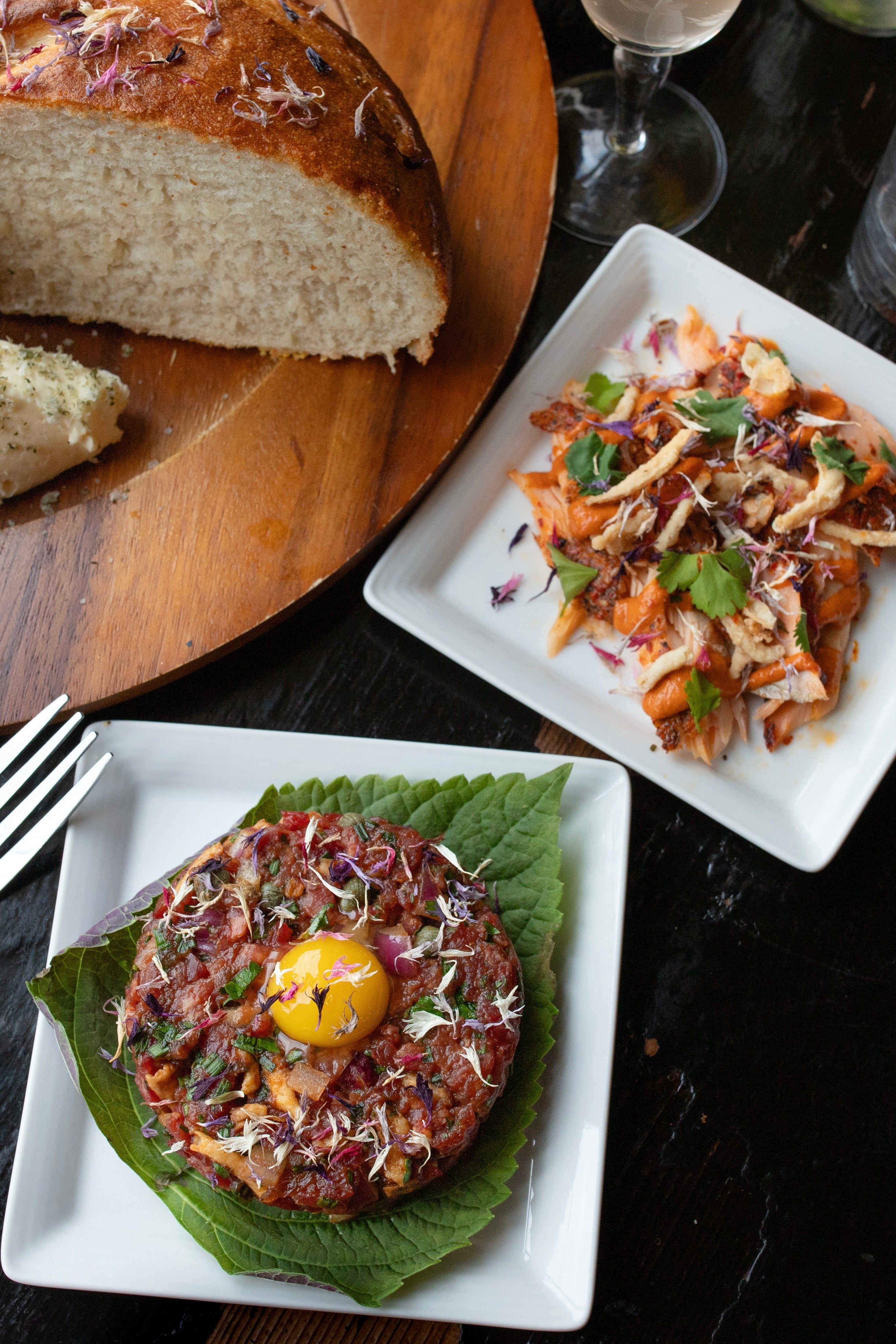 Photo of Vanissa Edible Flower Petals on Salmon tare tare. This is displayed on a green shiso leaf on square white plate with utensils and accompanying pieces. The photo is by chefs Mateja and Grills at Heritage Restaurant and Caviar Bar.