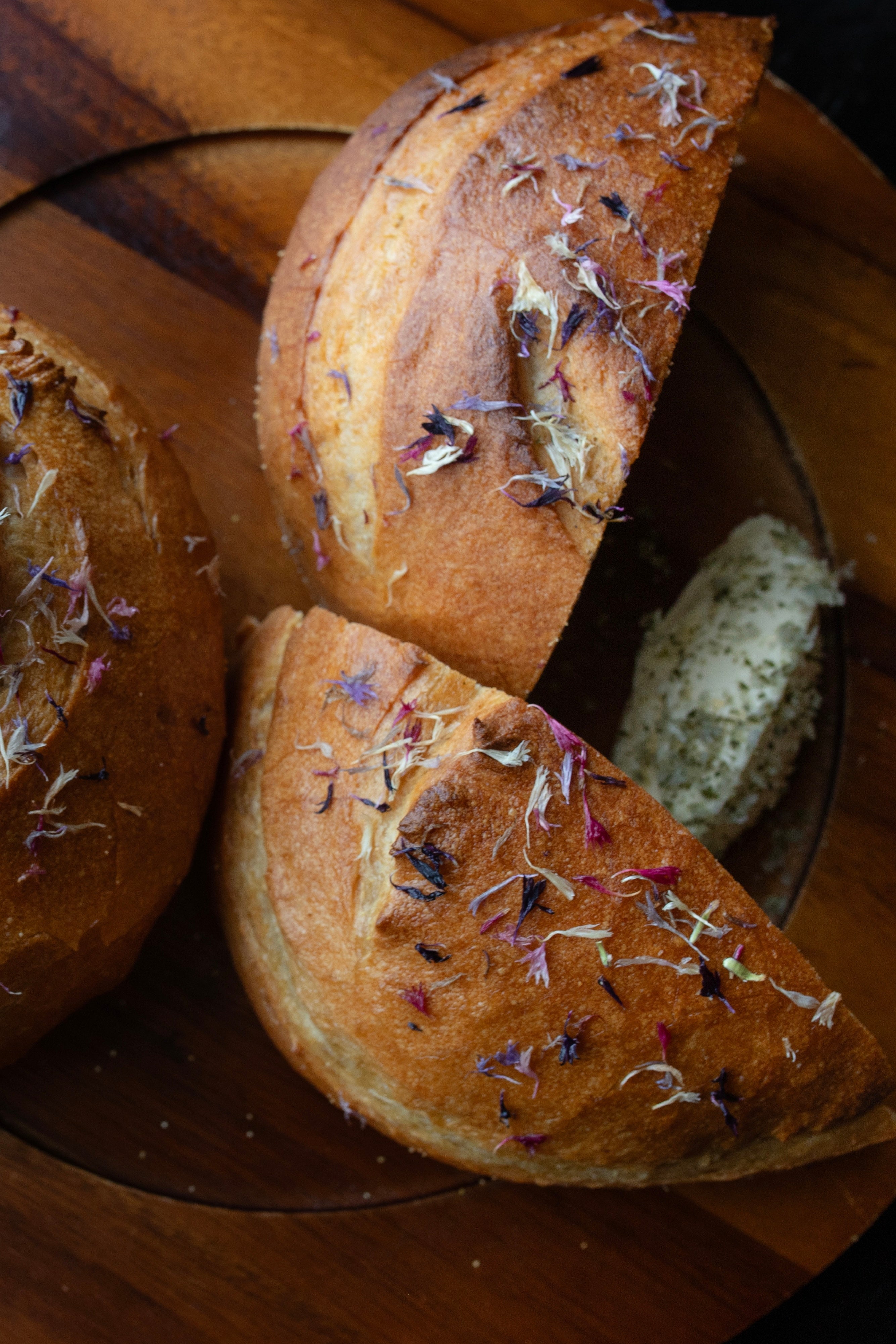 A photo of a halved round loaf of bread as well as a full round loaf. These are on a round wooden plate and topped with edible flower petals. The plate is on a black wood table and photographed from above looking down. The photo is by chefs Mateja and Grills at Heritage Restaurant and Caviar Bar.