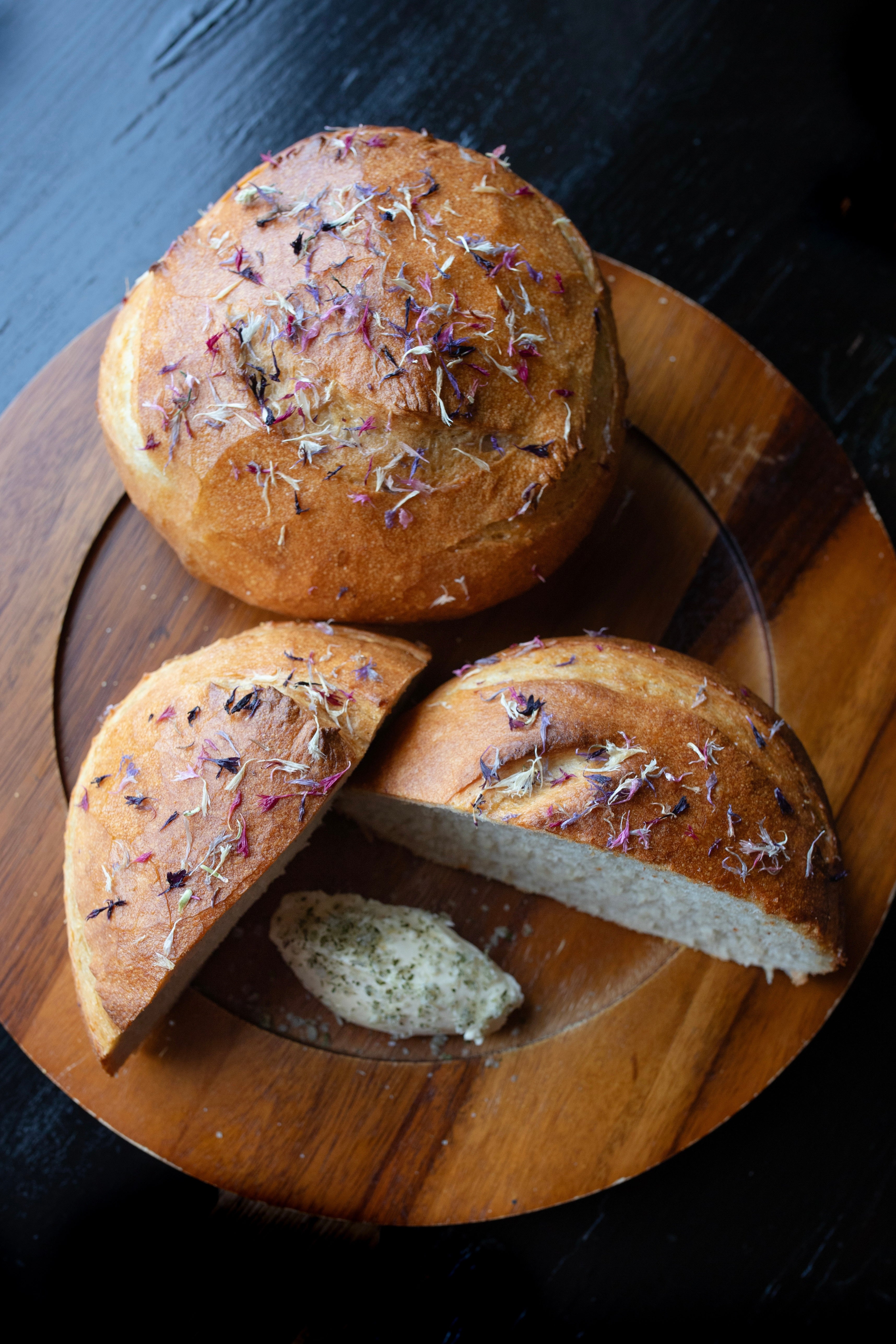 A photo of a halved round loaf of bread as well as a full round loaf. These are on a round wooden plate and topped with edible flower petals. The plate is on a black wood table and photographed from above looking down. The photo is by chefs Mateja and Grills at Heritage Restaurant and Caviar Bar.