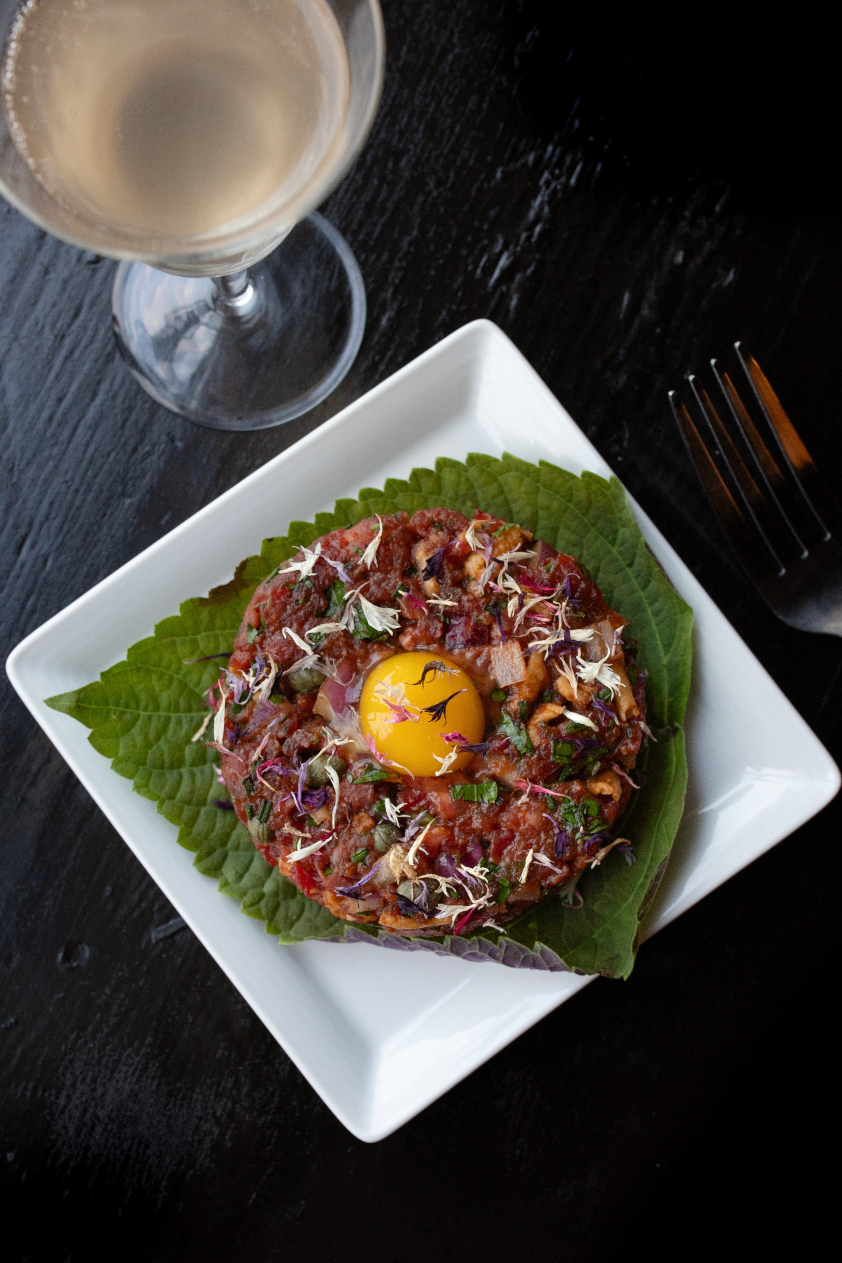 Photo of Vanissa Edible Flower Petals on Salmon tare tare. This is displayed on a green shiso leaf on square white plate with utensils and accompanying pieces. The photo is by chefs Mateja and Grills at Heritage Restaurant and Caviar Bar.