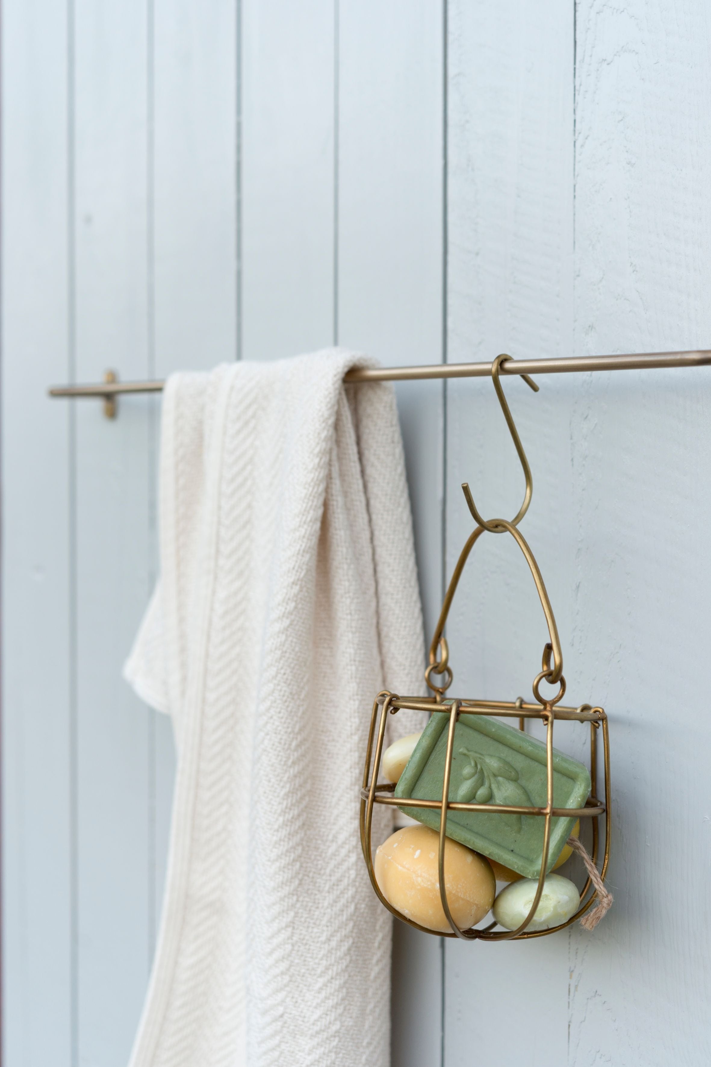 Small brass hook by Fog Linen Work in a bathroom on a brass towel holder holding brass wire jar of soaps