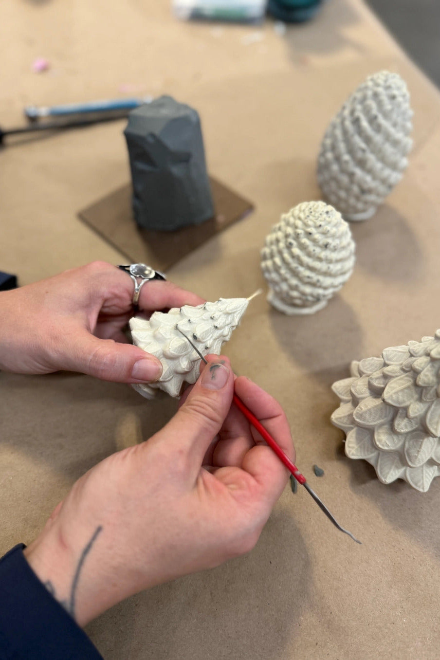 A close-up of a person's hands carving a small white textured pinecone sculpture with a red-handled tool on a tan workbench, with other finished pinecone sculptures in the background. ©Mo&Co.