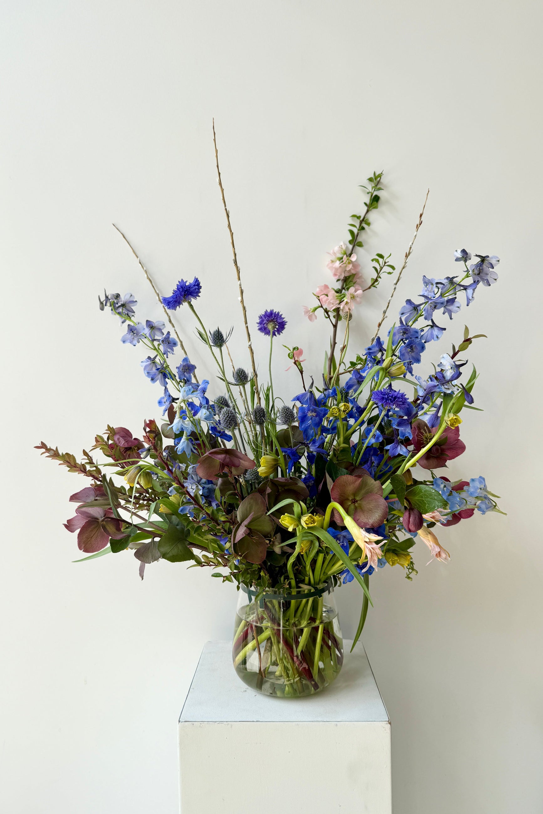 Vase of deep blueand purple flowers flowers featuring delphinium and Hellebore on a white pedestal against a white background ©Sprout Home #size_$130