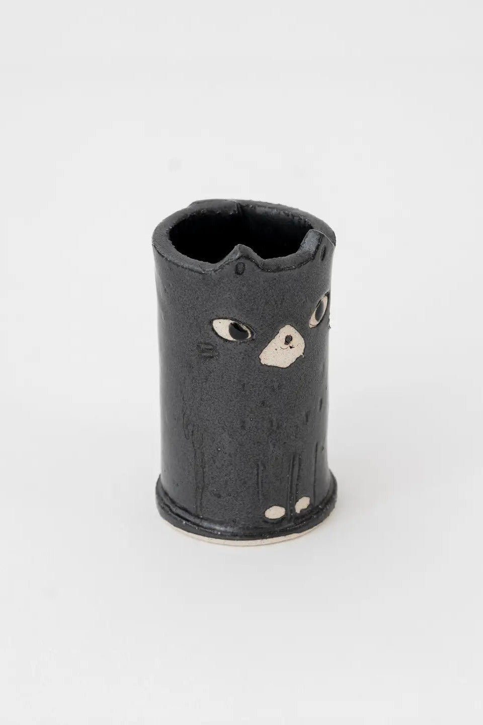 A small black ceramic cylindrical container with a stylized cat face painted on the front, photographed against a plain light gray background. ©Great Zakka