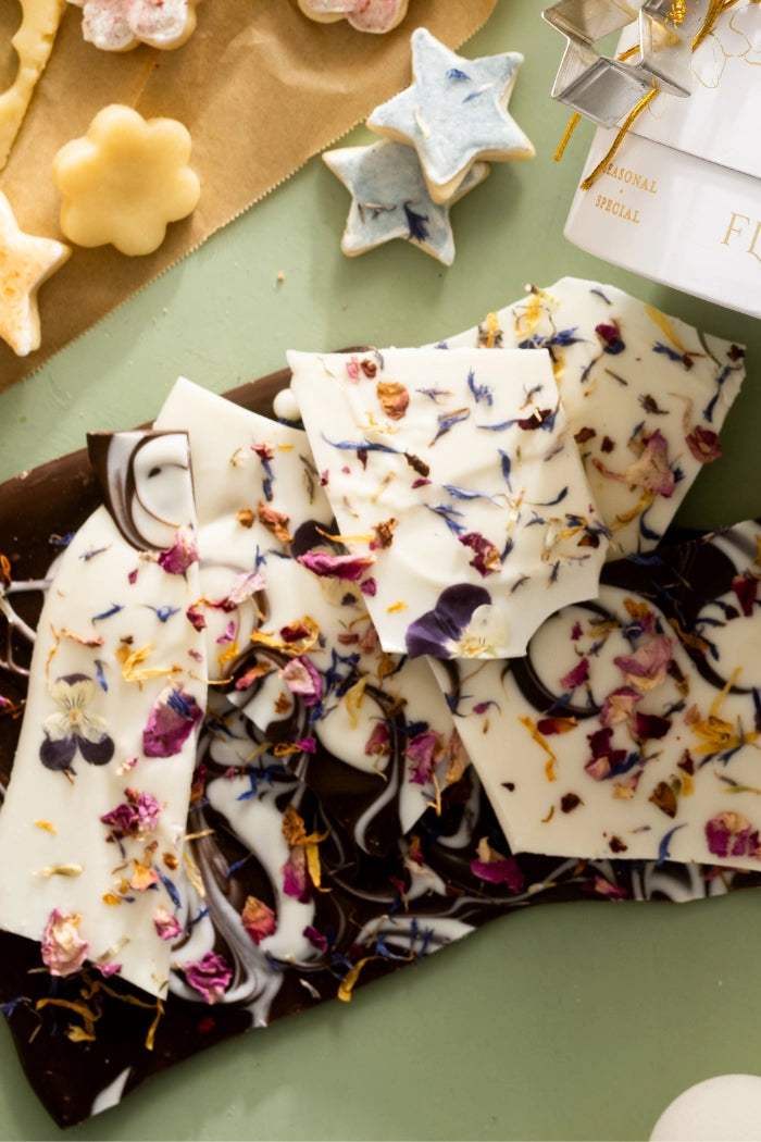 Decorative chocolate bark with floral toppings on a green surface, next to cookie cutters and a box labeled 'Flowercru'. ©Flouwer Co.