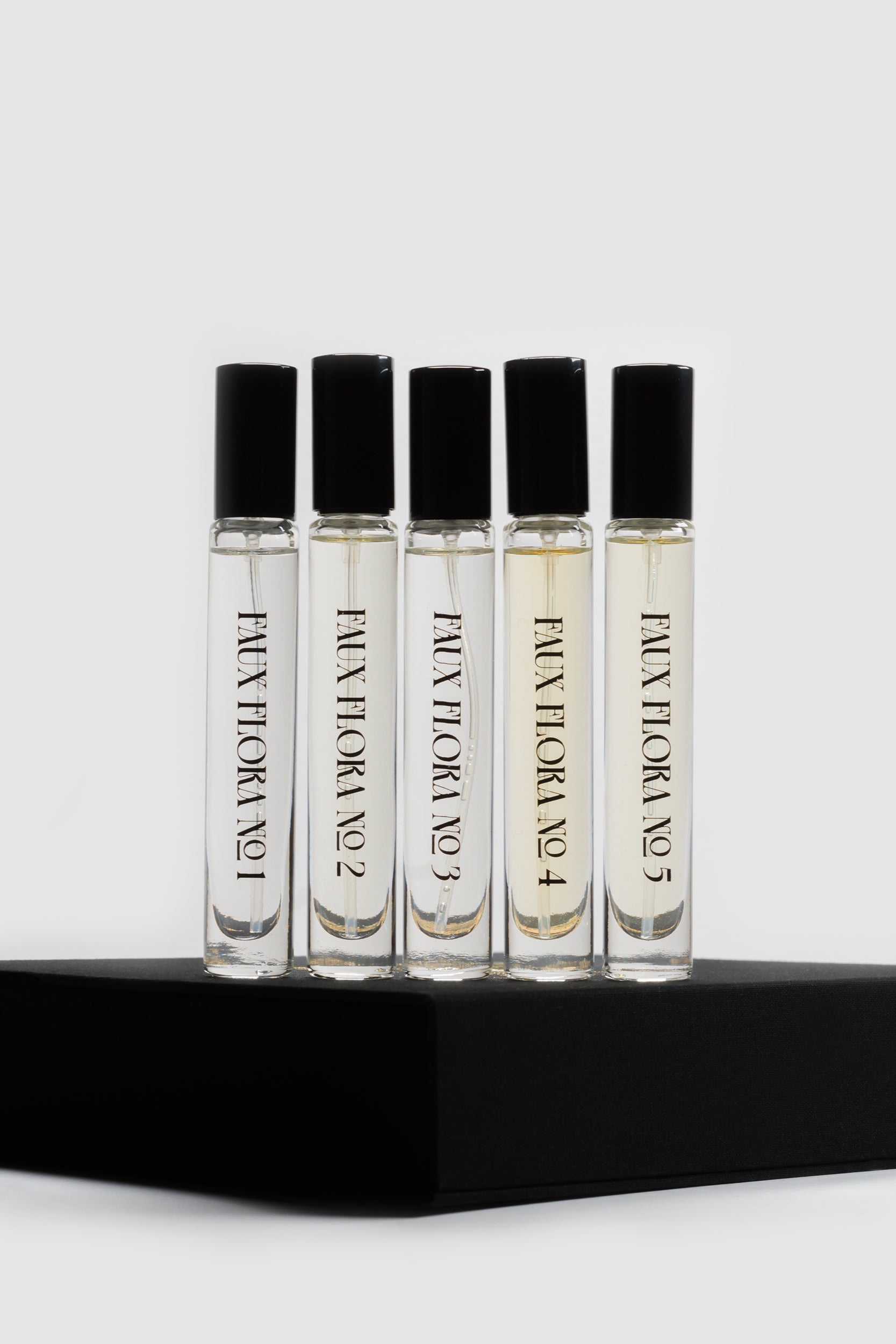 Set of five small perfume bottles with black caps of Faux Floral Discovery Set on a black platform against a light gray background ©Fischersund