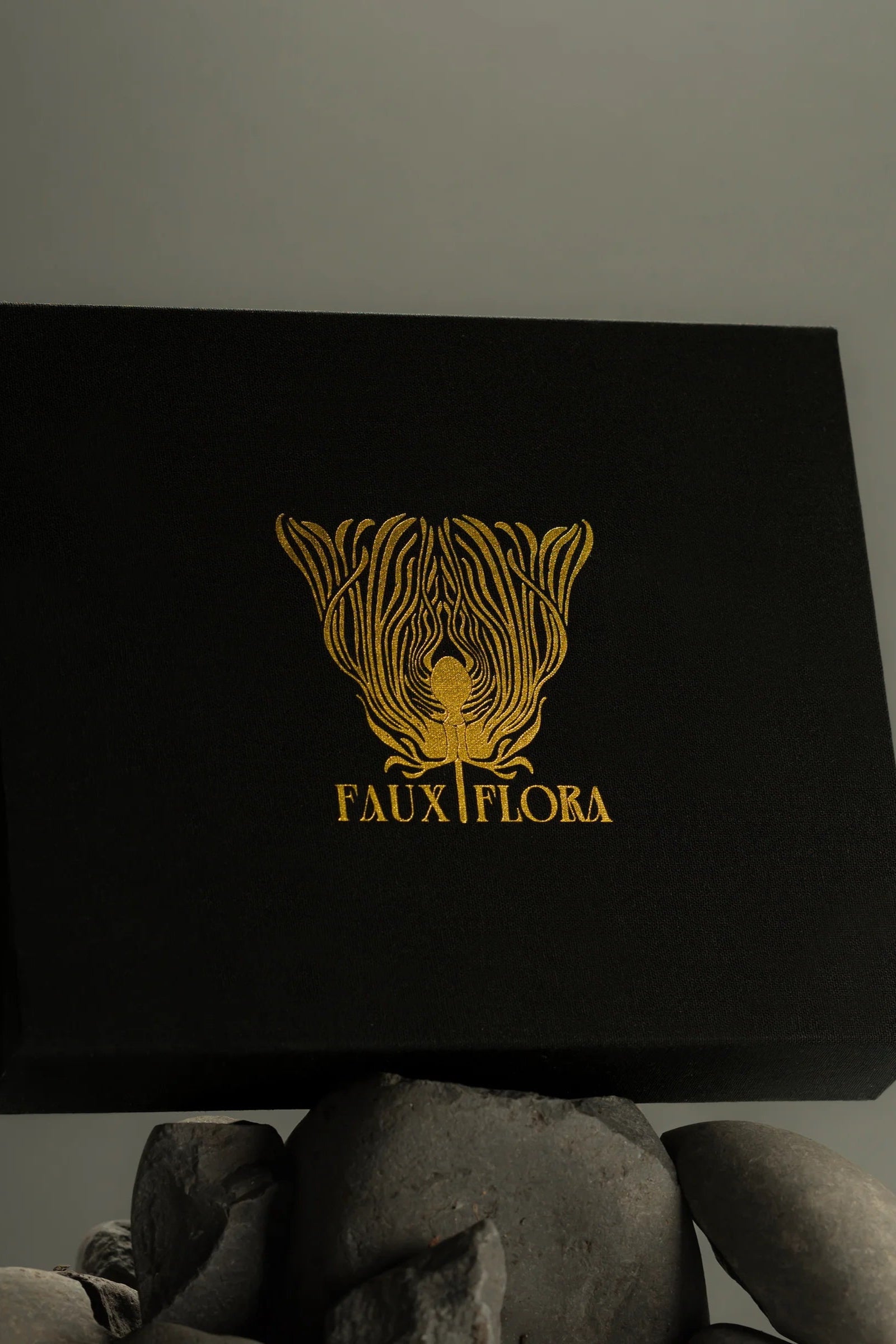 Black box with gold 'FAUXFLORA' logo held by rocks against a gray background ©Fischersund