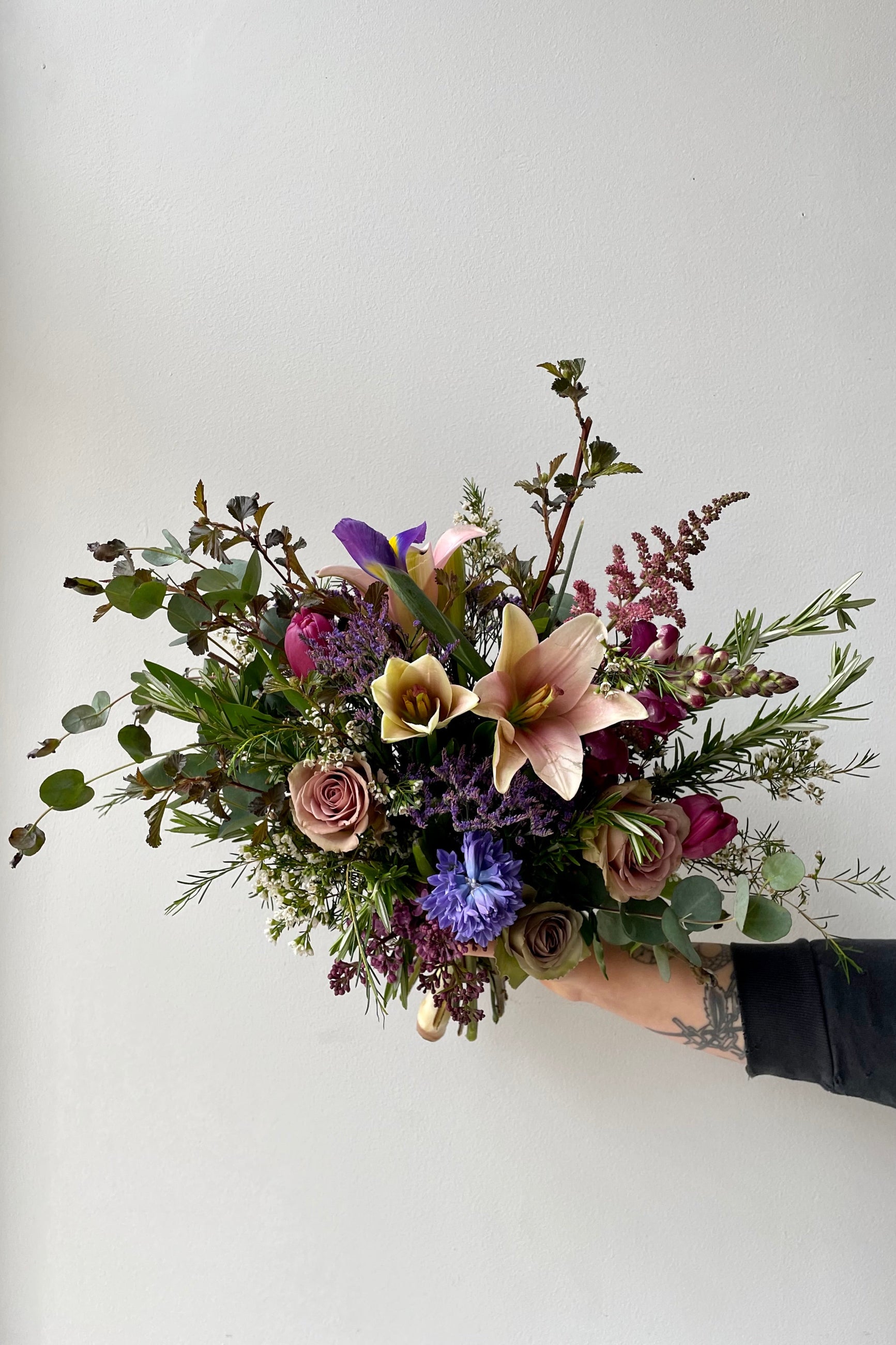 A mid May custom storm arrangement with Lillies and purple roses by Sprout Home being held against a white wall. ©Sprout Home #size_$130