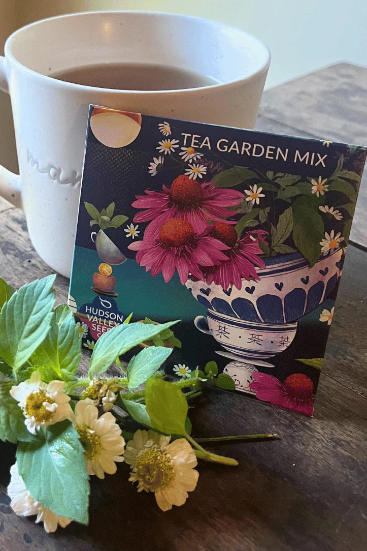 White mug with tea and a tea bag labeled 'Tea Garden Mix' on a wooden surface with flowers. ©Hudson Valley Seed Co.