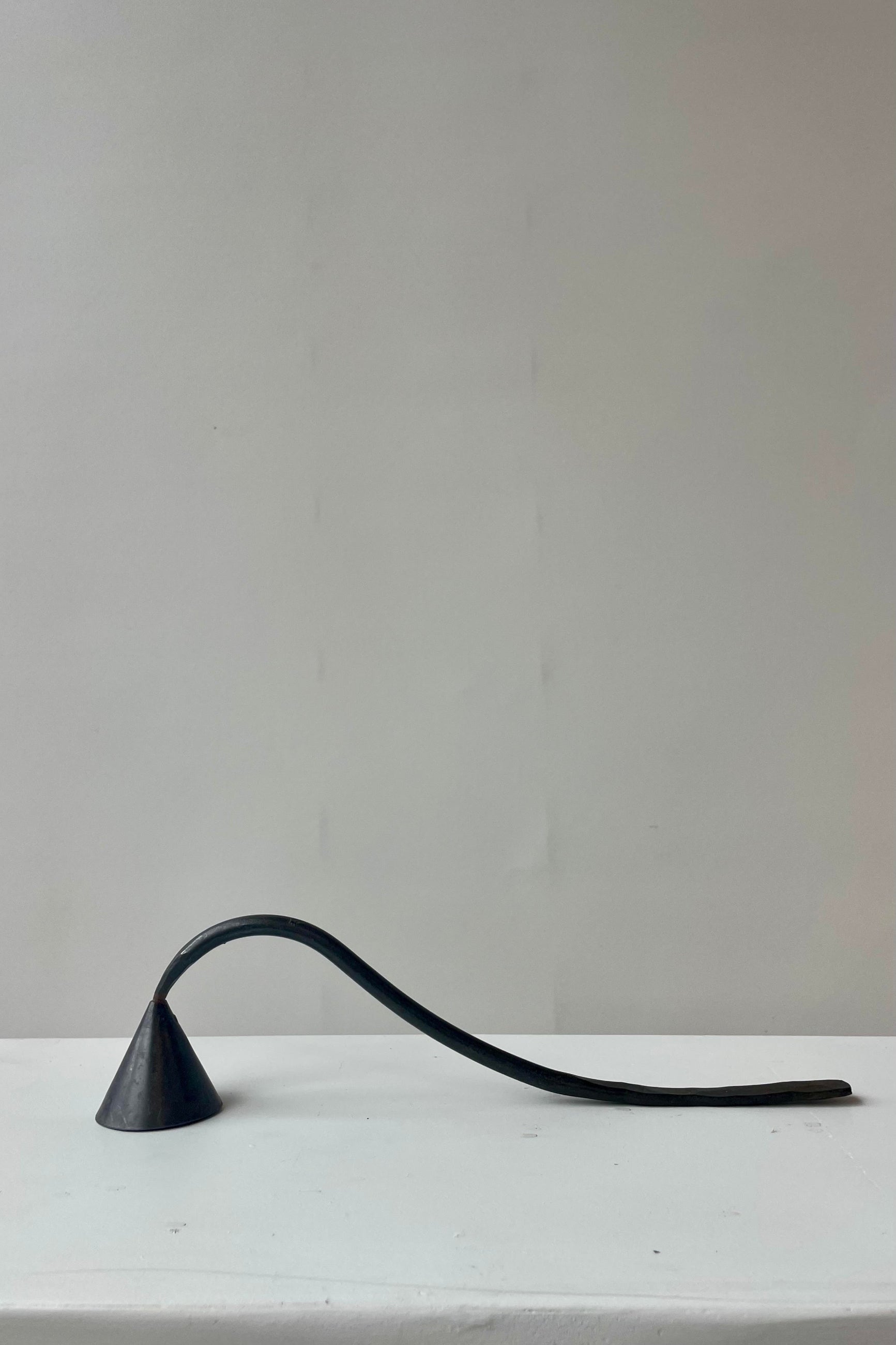 Photo of a black metal candle snuffer on a white surface against a white wall. The handle curves up and then down to rest on the same surface. ©Sprout Home