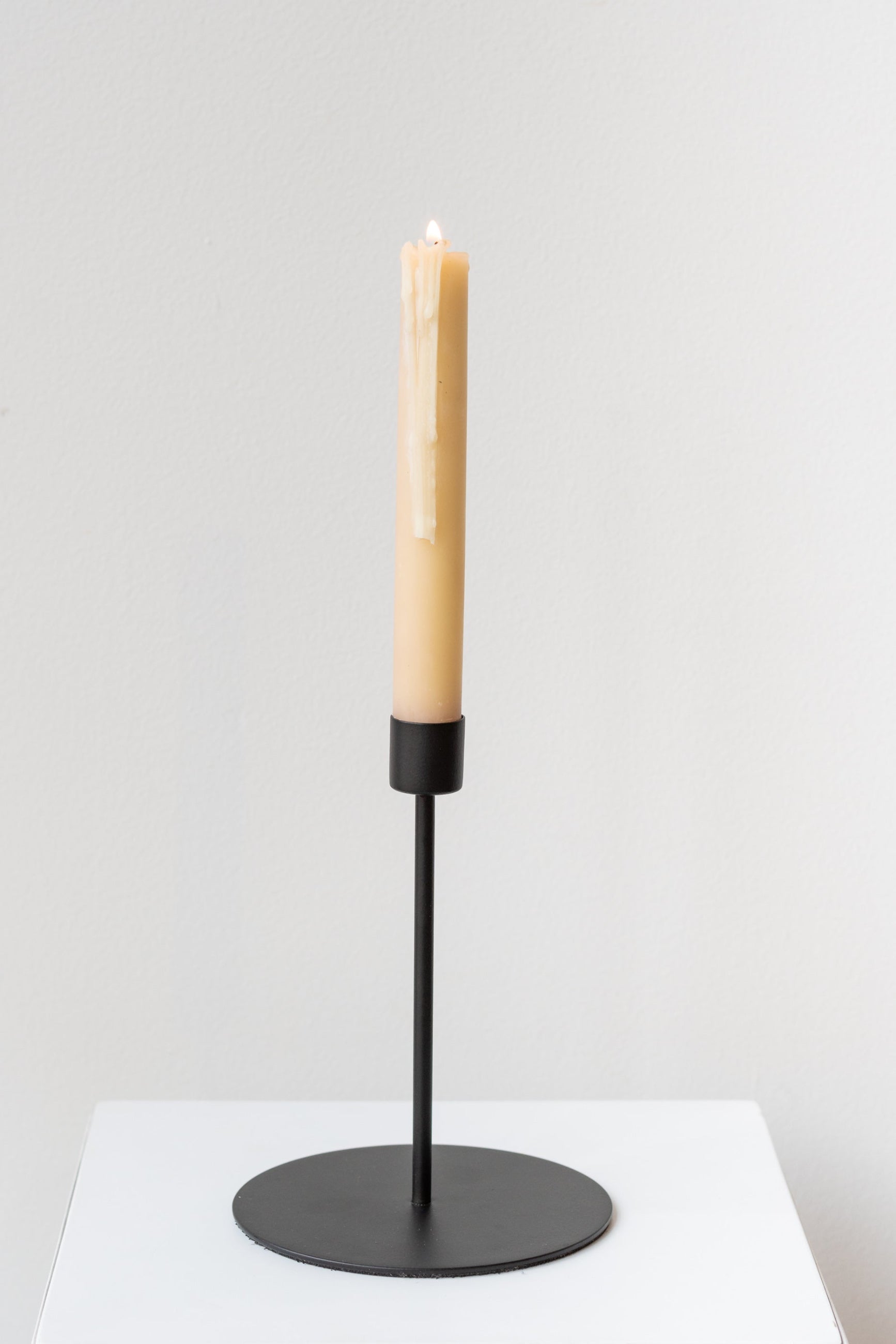 Black tall taper candle holder on a white surface in a white room - inside the candle holder is a lit beige taper candle ©Sprout Home