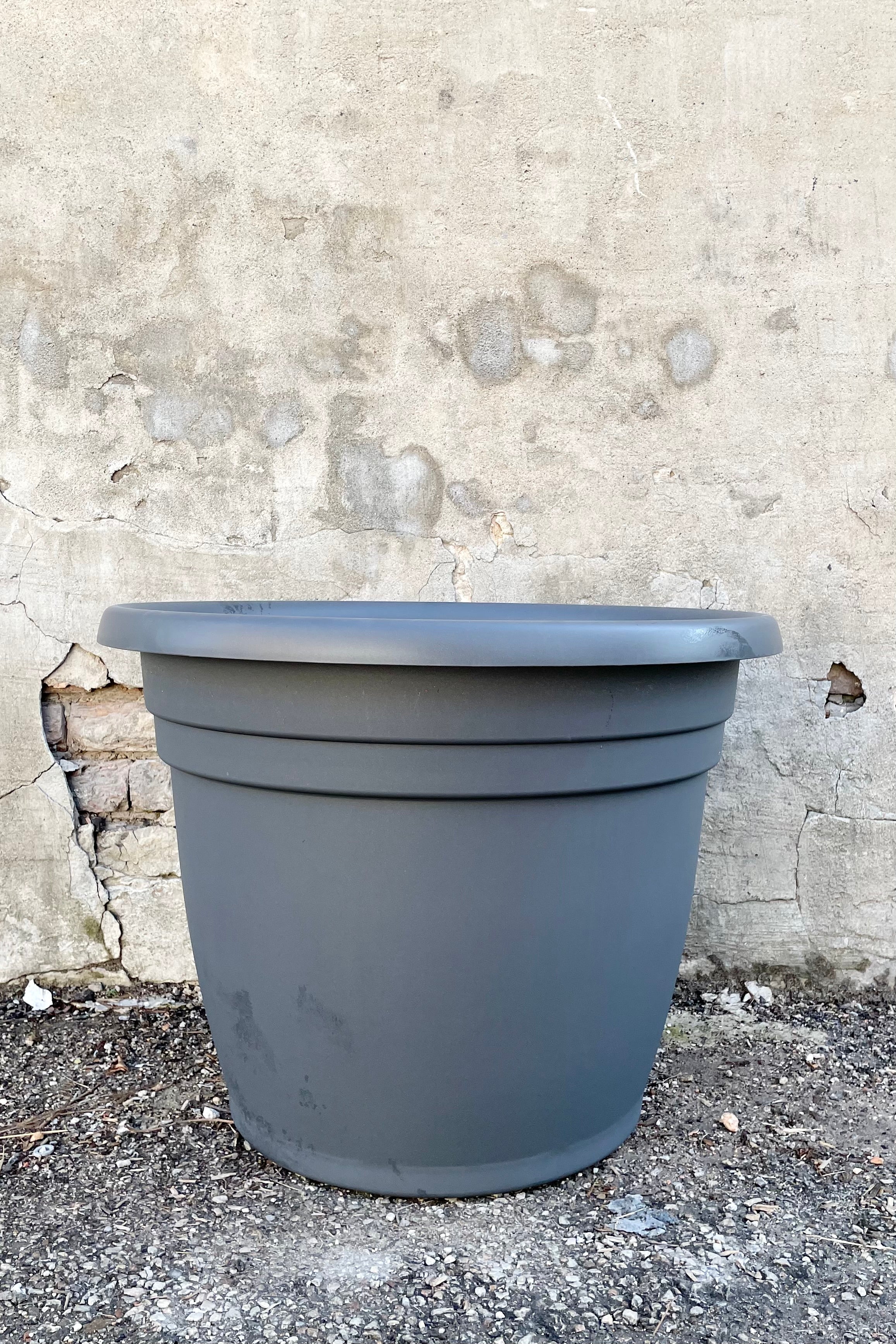 The emma round charcoal 25" planter against a concrete wall.