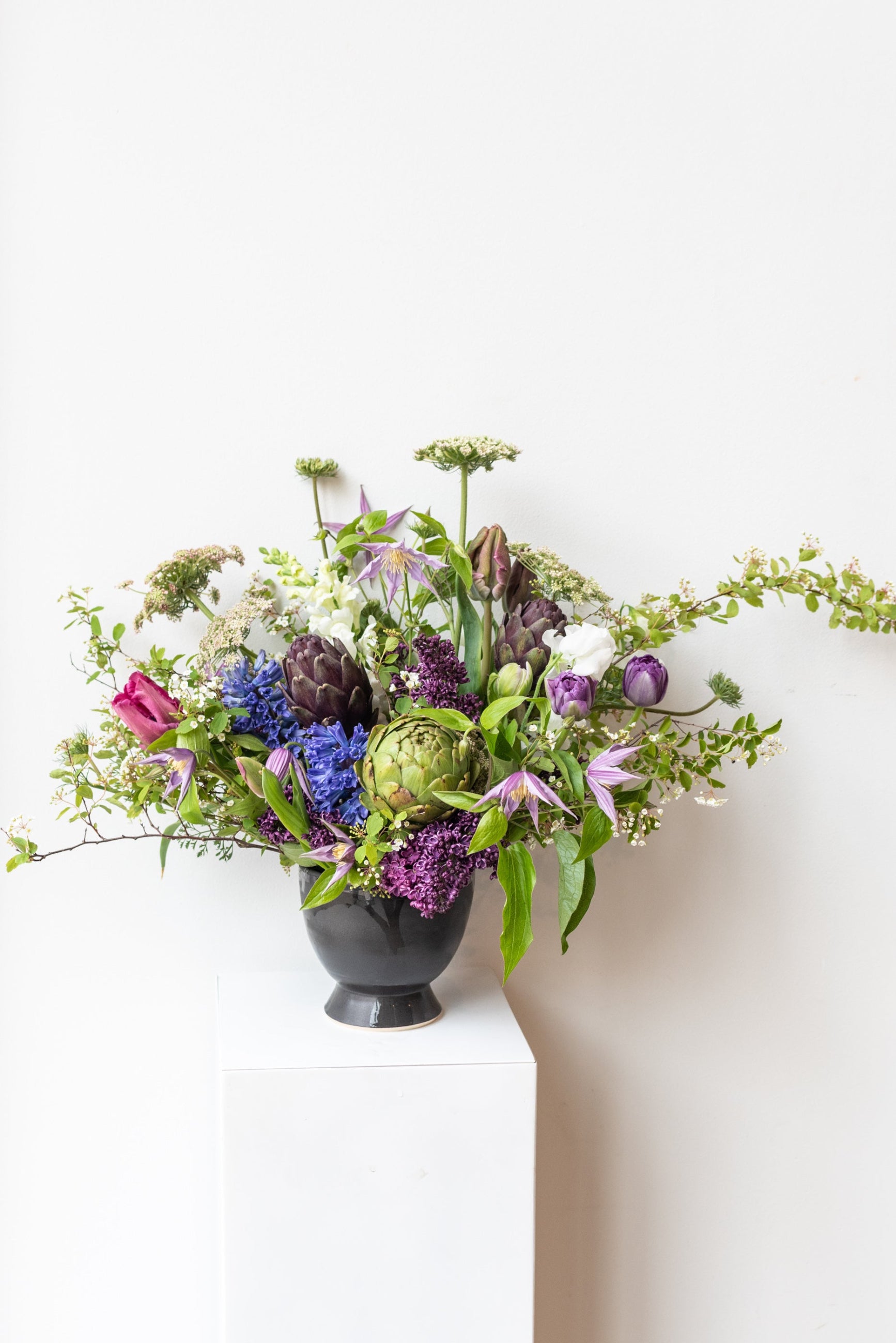 Large storm floral arrangement by Sprout Home sits in a black Little Fire Ceramics compote on a white pedestal in a white room ©Sprout Home #size_$200