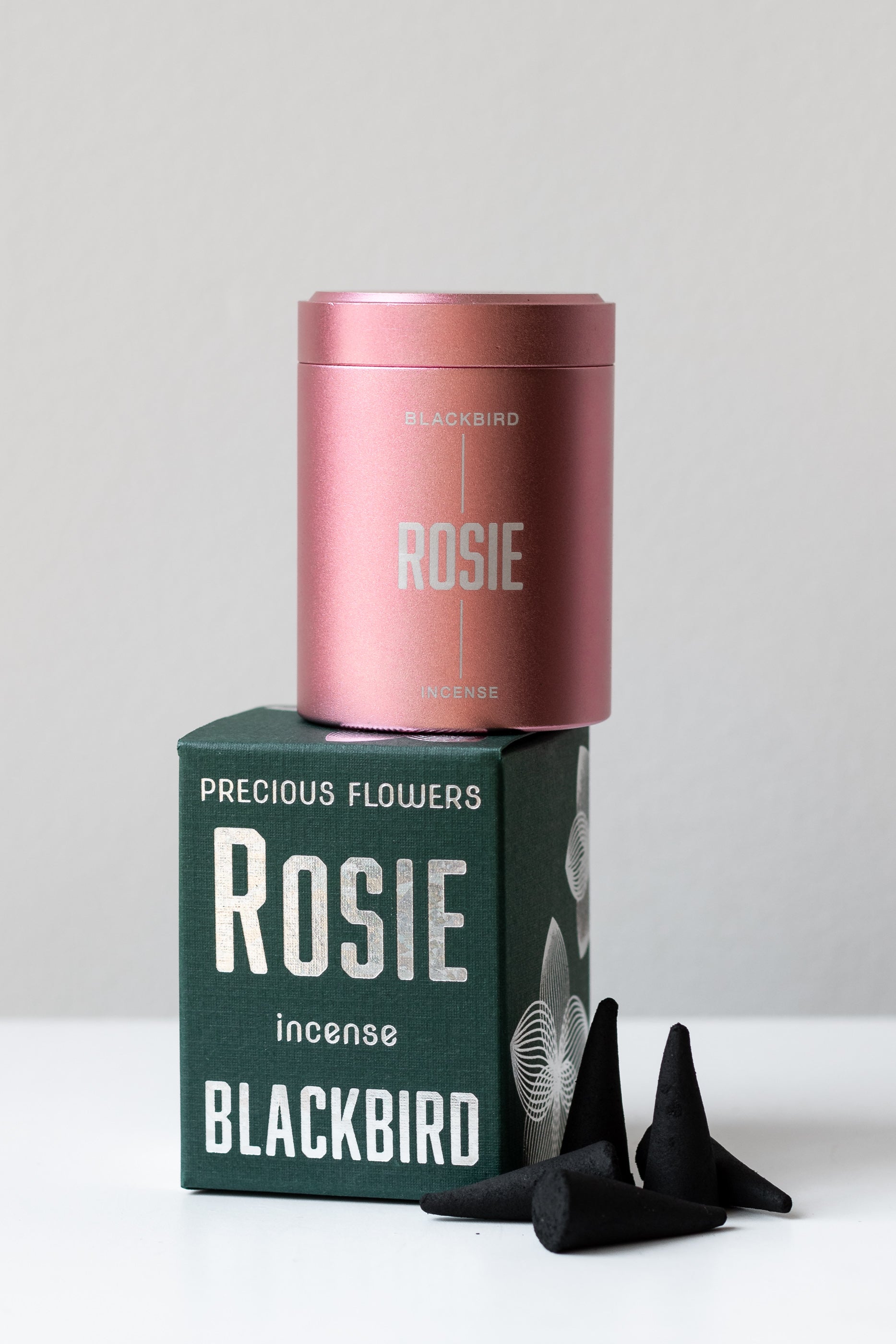Rosie incense by BLACKBIRD sits on a white surface in a white room. The pink incense tin is on top of the box, and five black incense cones are scattered to the side of the box.