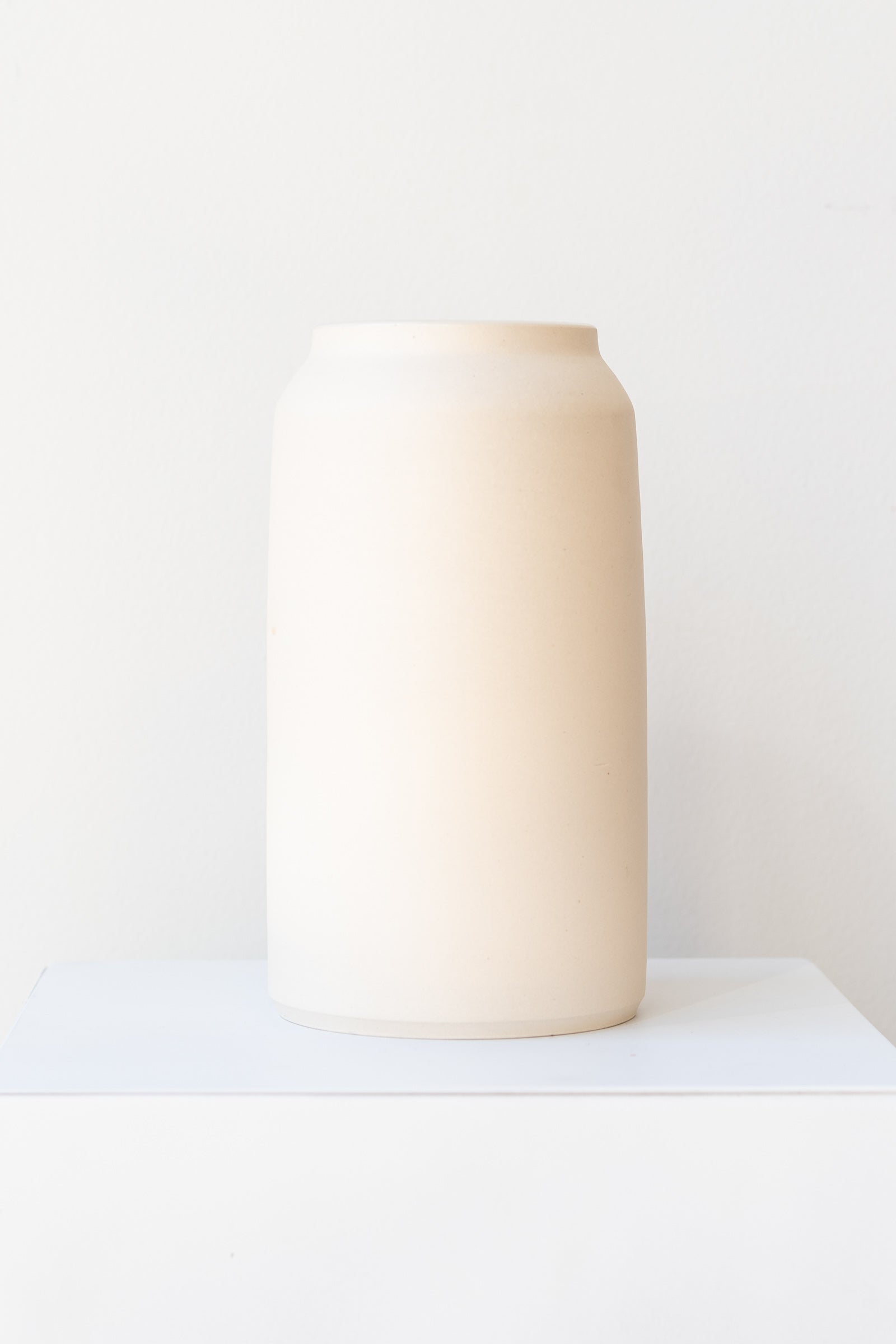One cream colored bouquet vase sits on a white surface in a white room. The vase is tall and cylindrical, with a slight rim at the top. It is photographed straight on.