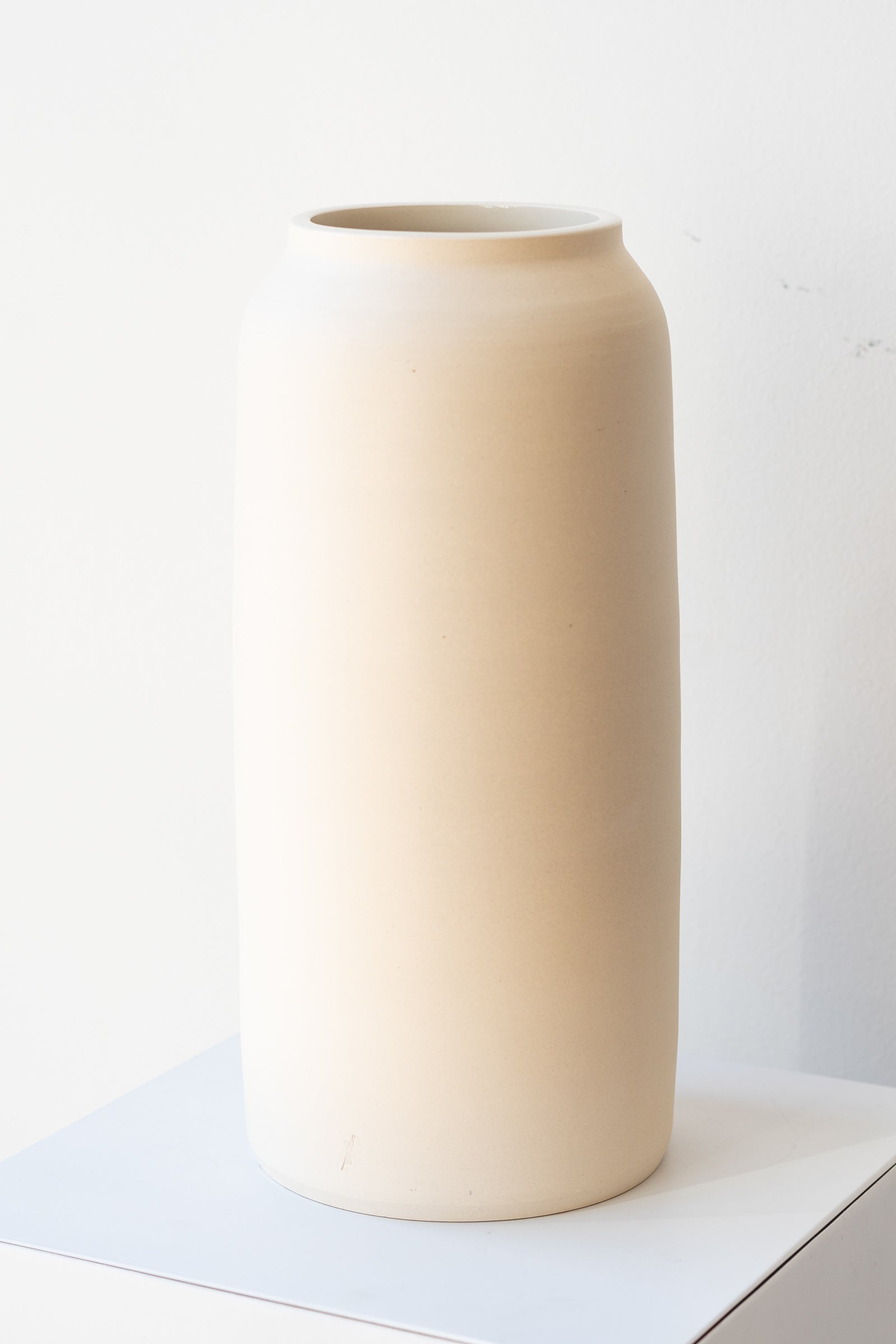 One cream colored bouquet vase sits on a white surface in a white room. The vase is tall and cylindrical, with a slight rim at the top. It is photographed at a slight angle to show the clay detail.
