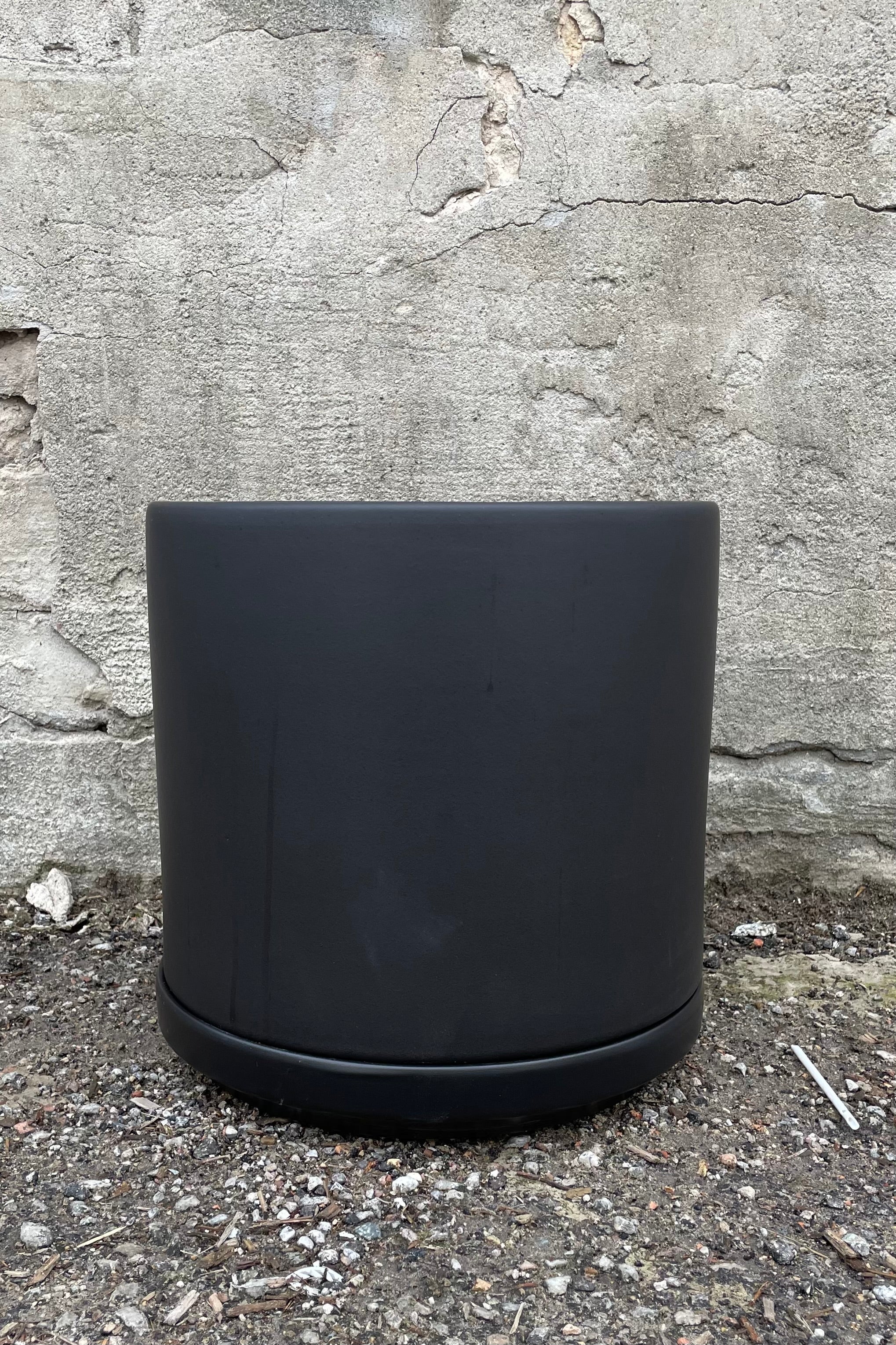 The 10" solid cylinder and saucer in black against a concrete wall.
