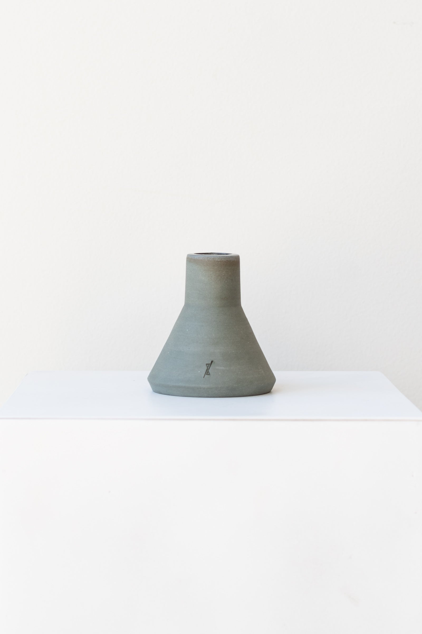 One triangular grey stoneware candle holder sits on a white, flat surface in a white room. It has a small logo etched into the clay near the base. It is photographed straight on.