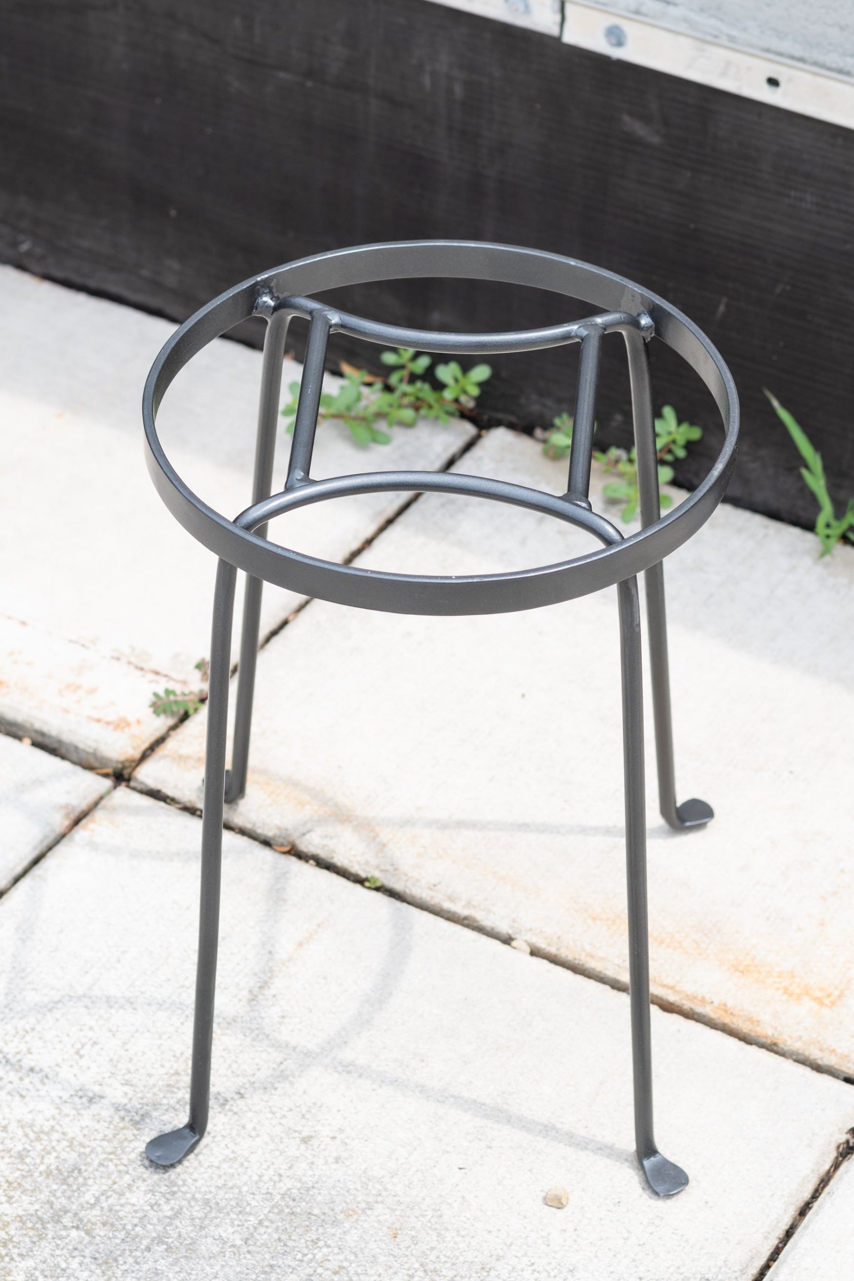 A wrought iron plant stand with a round top and three legs, displayed on a stone surface. ©Sprout Home