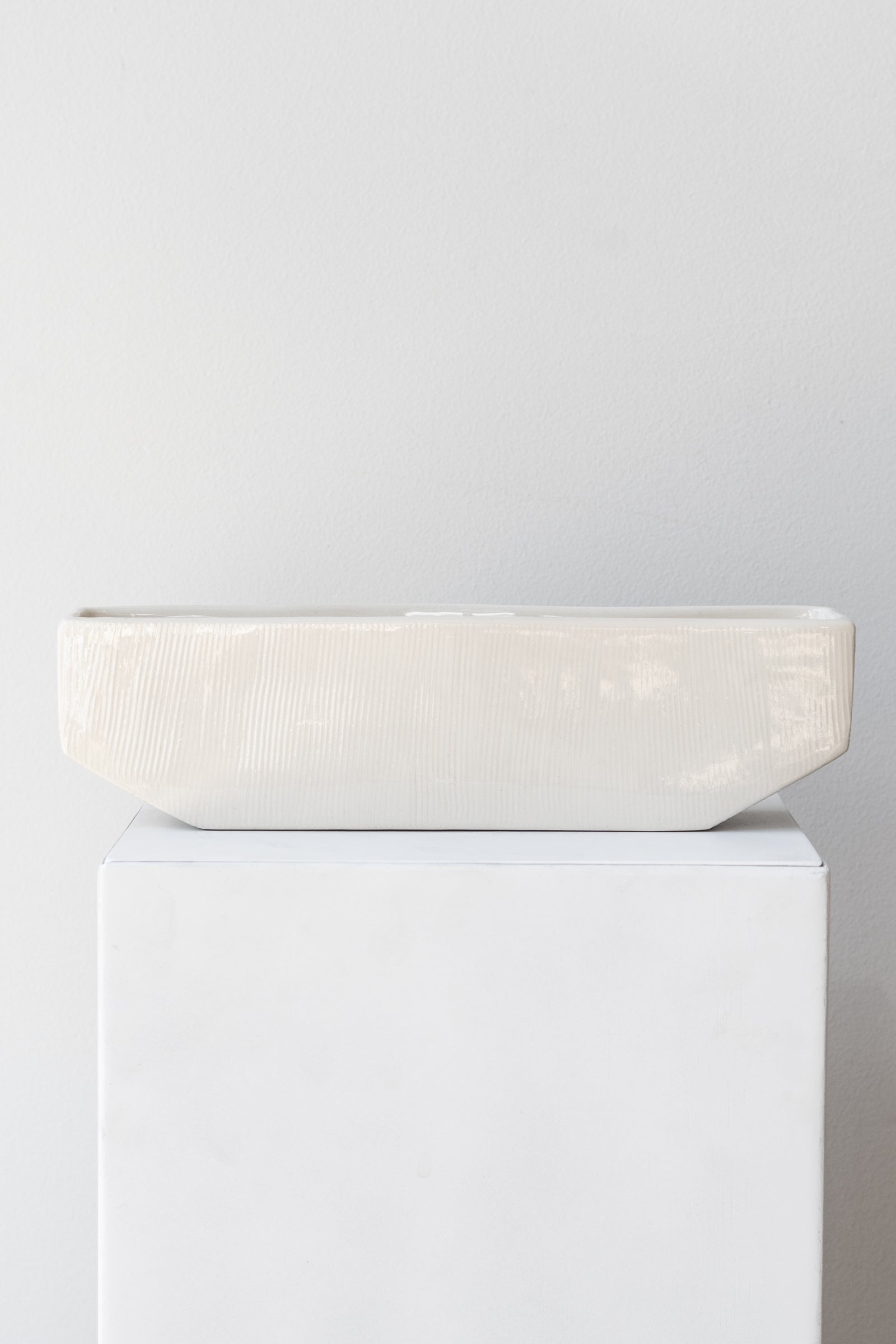 One medium rectangular white planter sits on a white square pedestal in a white room. The planter is empty. It is photographed straight on.