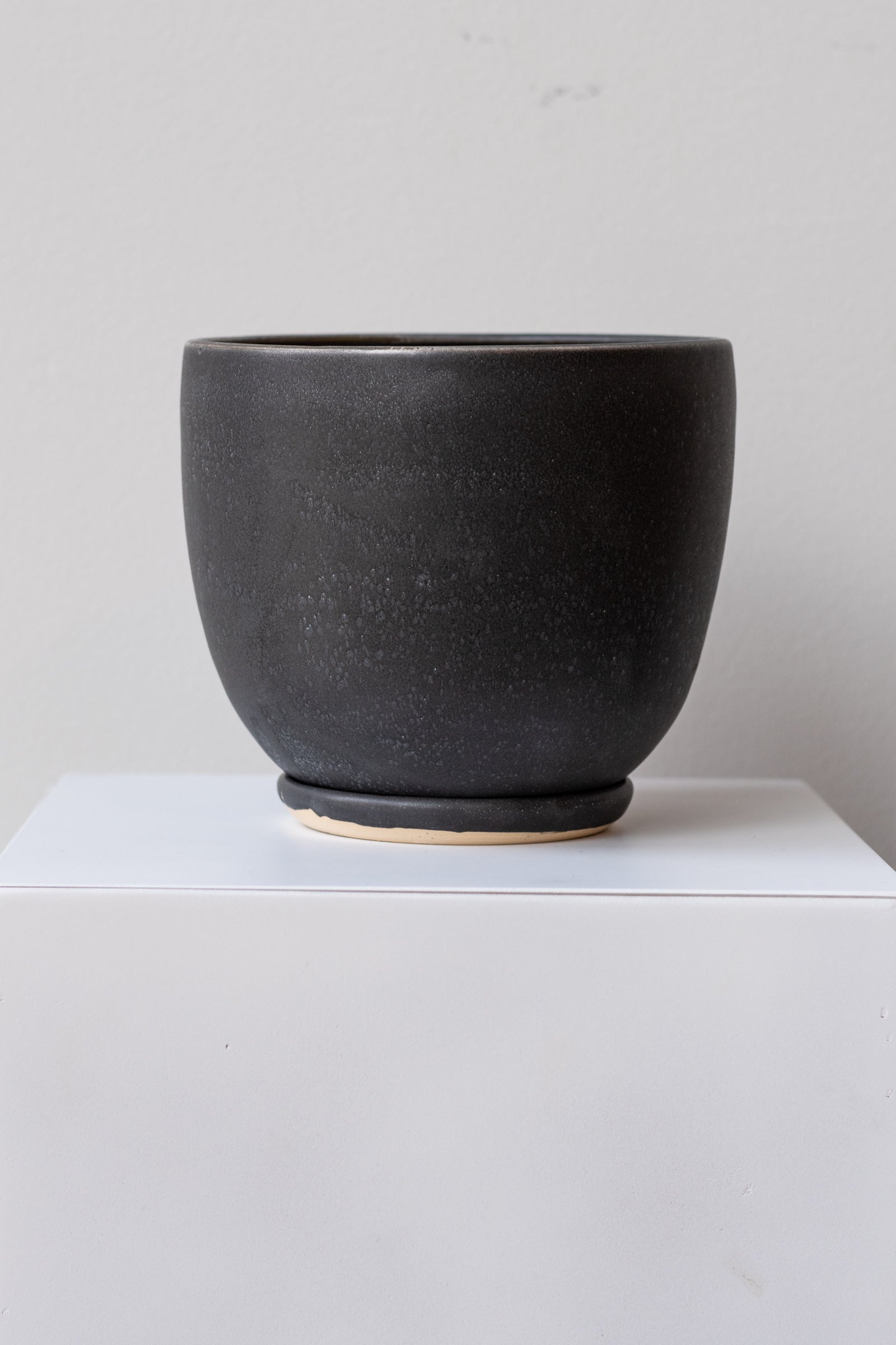 The planter is glazed black with white speckles. It sits on a small drainage tray. The planter is empty. It is photographed straight on.