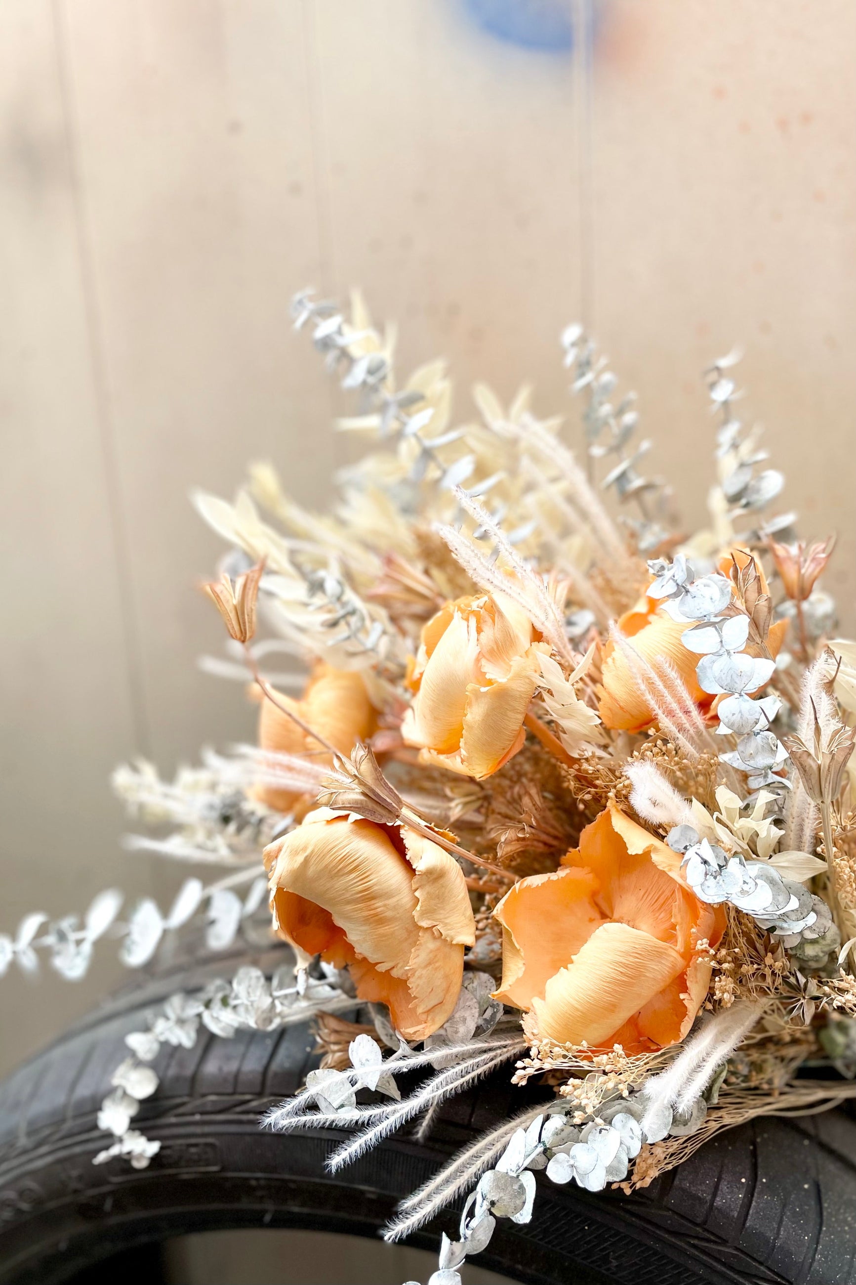 The $125 option of Preserved Arrangement Creamsicle by Sprout Home lies on a tire against a brown fence ©Sprout Home #size_$125