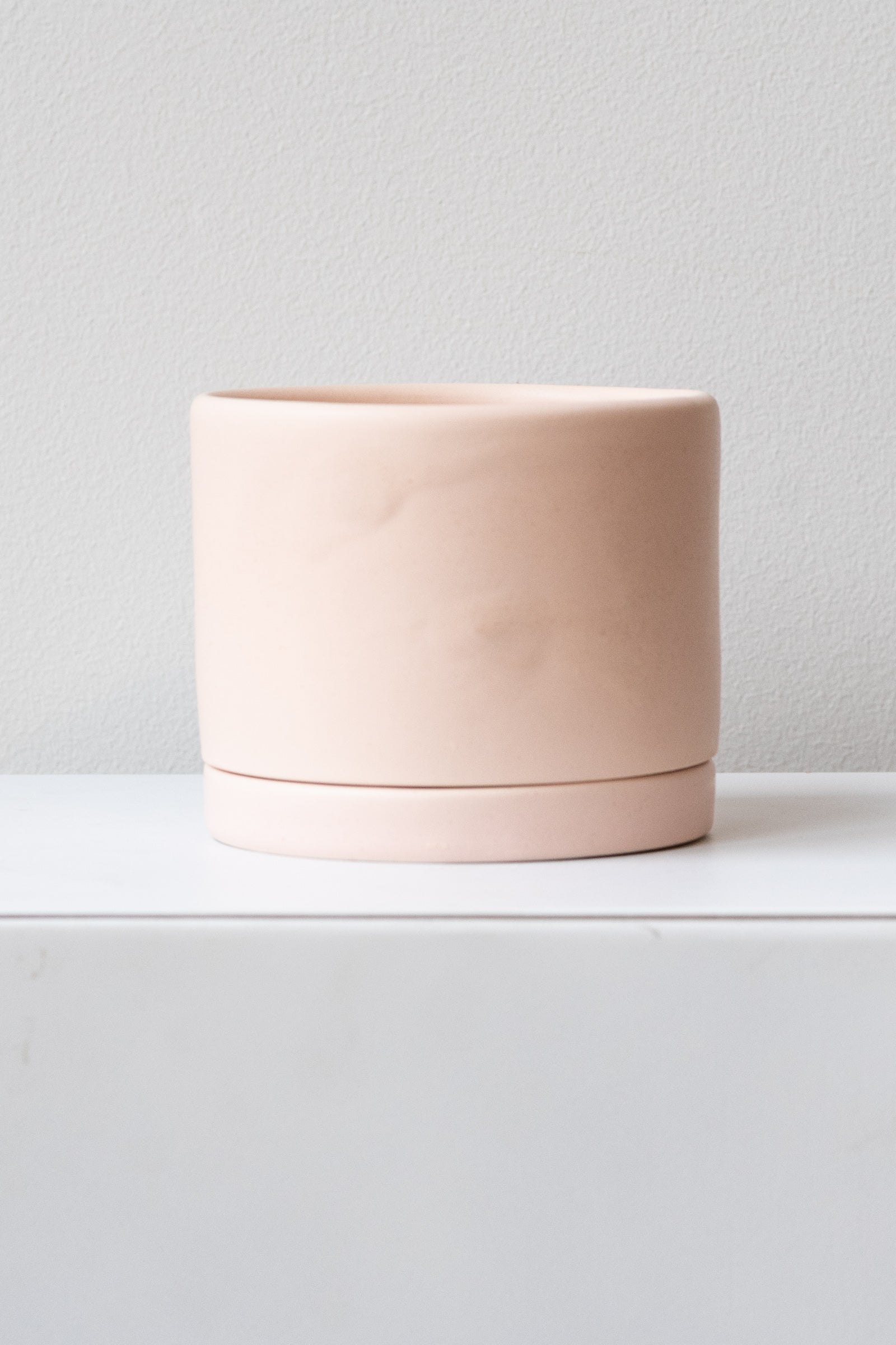 A medium pink ceramic planter sits on a white surface in a white room. The planter has a matching drainage tray. The planter is empty. It is photographed straight on.