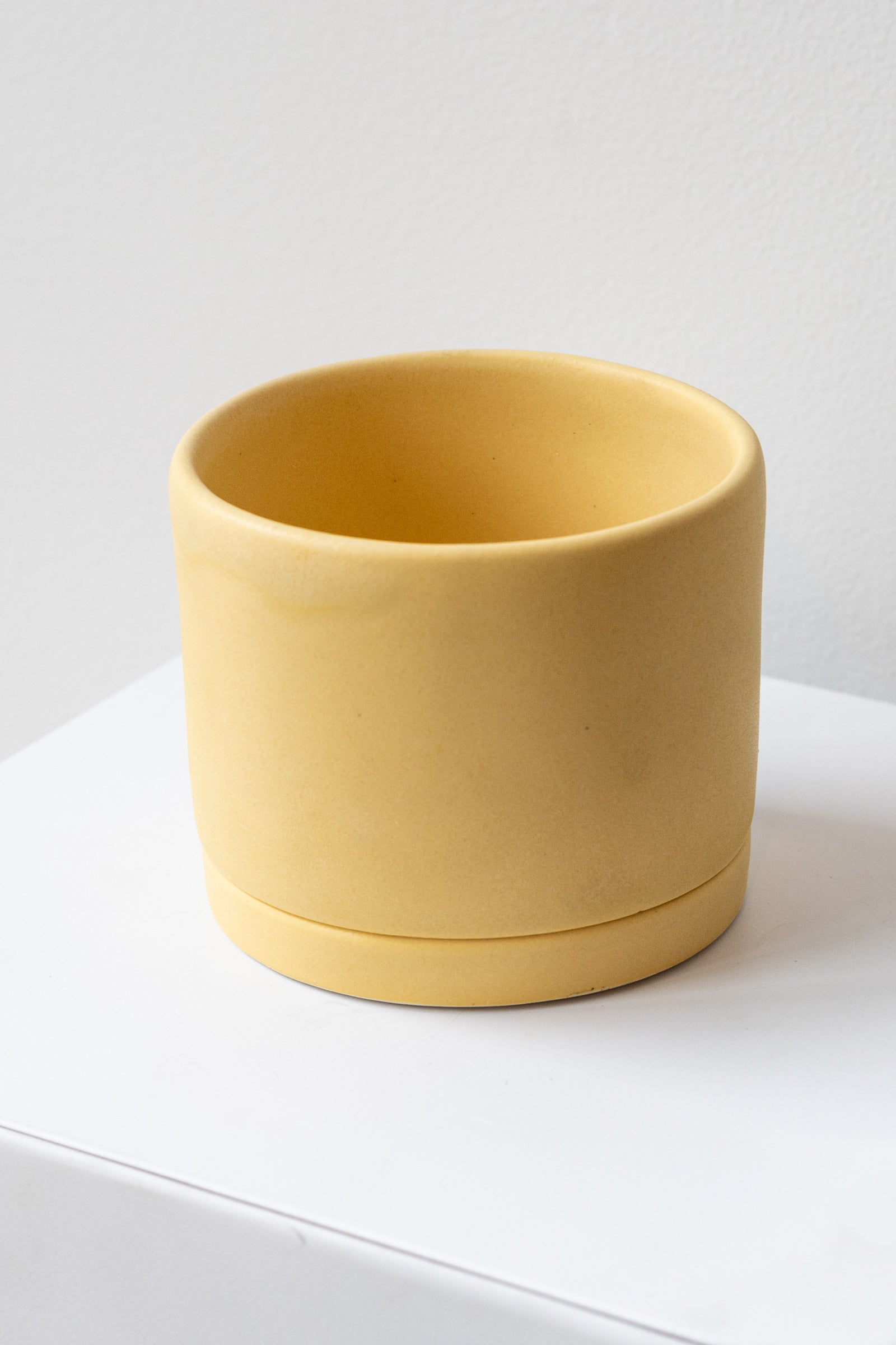 A medium yellow ceramic planter sits on a white surface in a white room. The planter has a matching drainage tray. The planter is empty. It is photographed closer and at an angle.