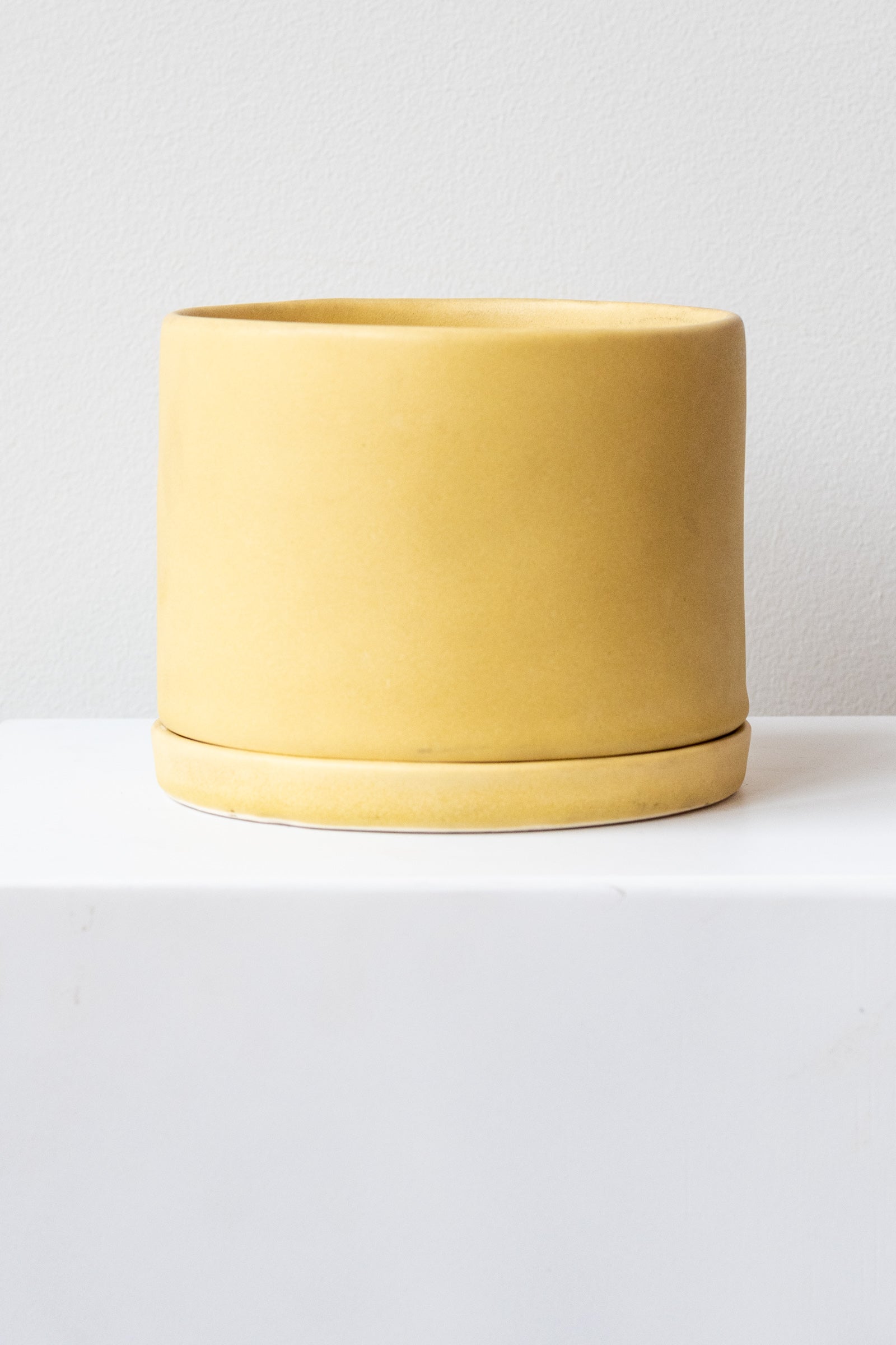 A large yellow ceramic planter sits on a white surface in a white room. The planter has a matching drainage tray. The planter is empty. It is photographed straight on.