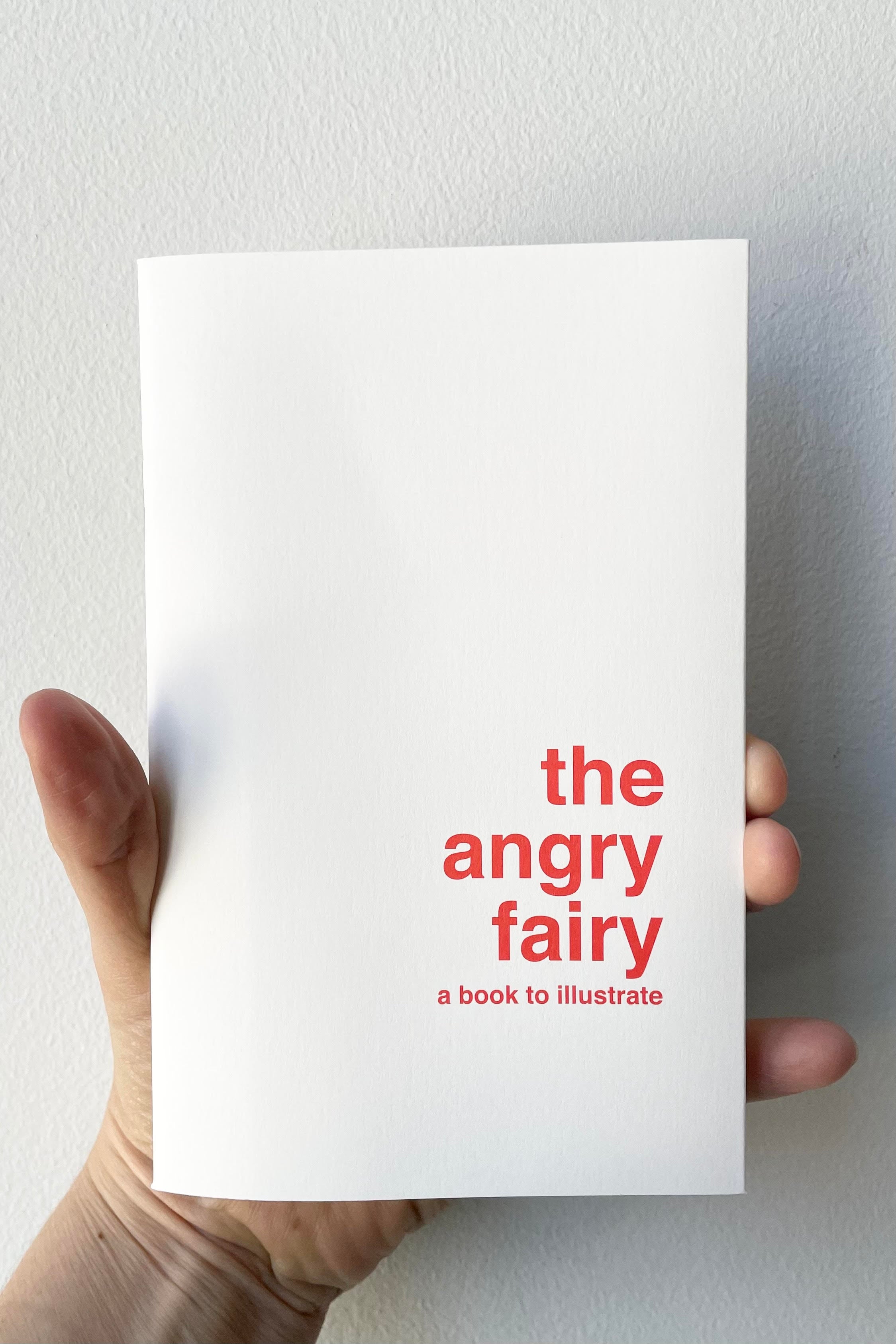 A hand holds The Angry Fairy - Book to Illustrate against a white backdrop.