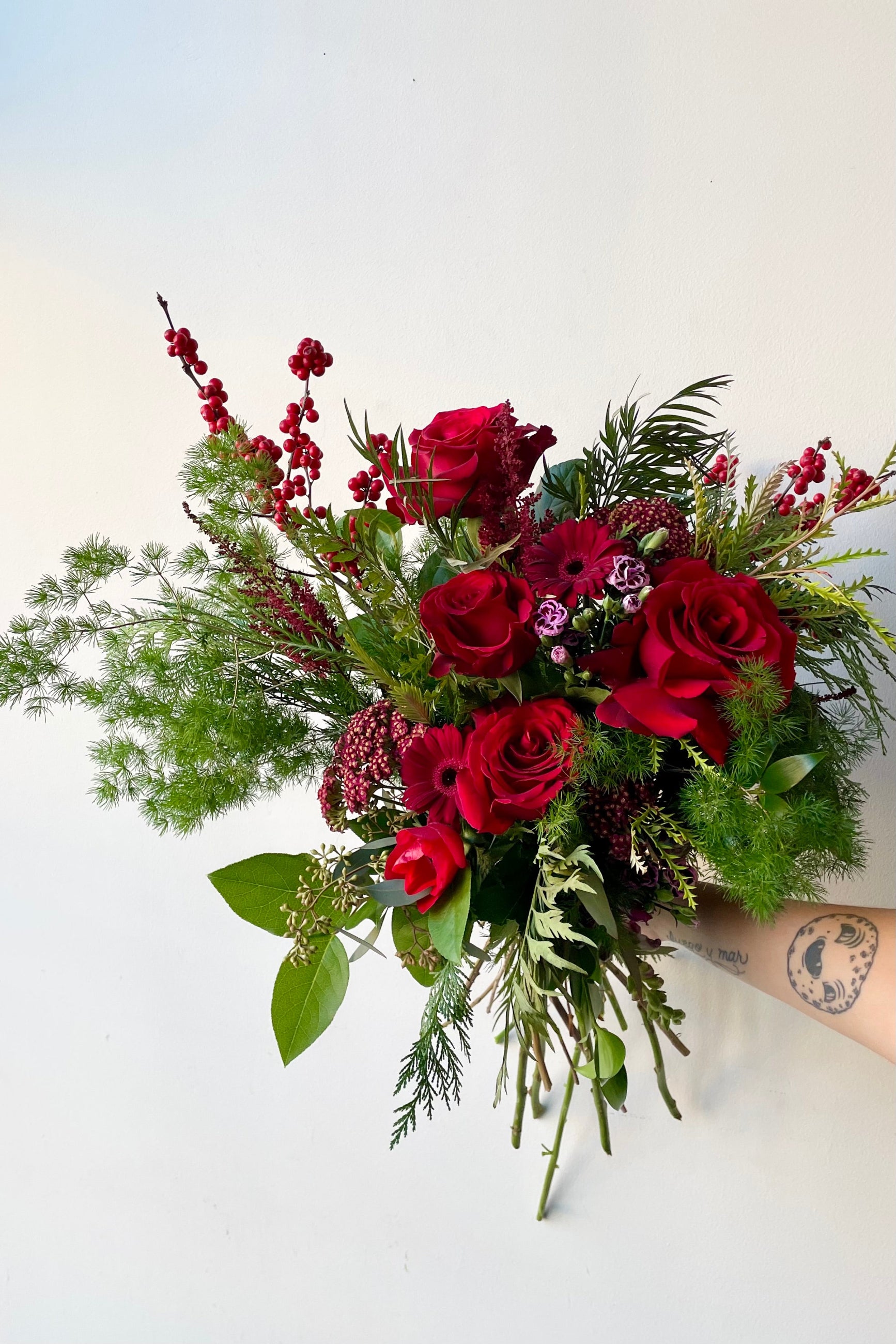 A hand holds an example of fresh Floral Arrangement Solstice with asparagus fern greens and reds from Sprout Home Floral in Chicago #size_$160