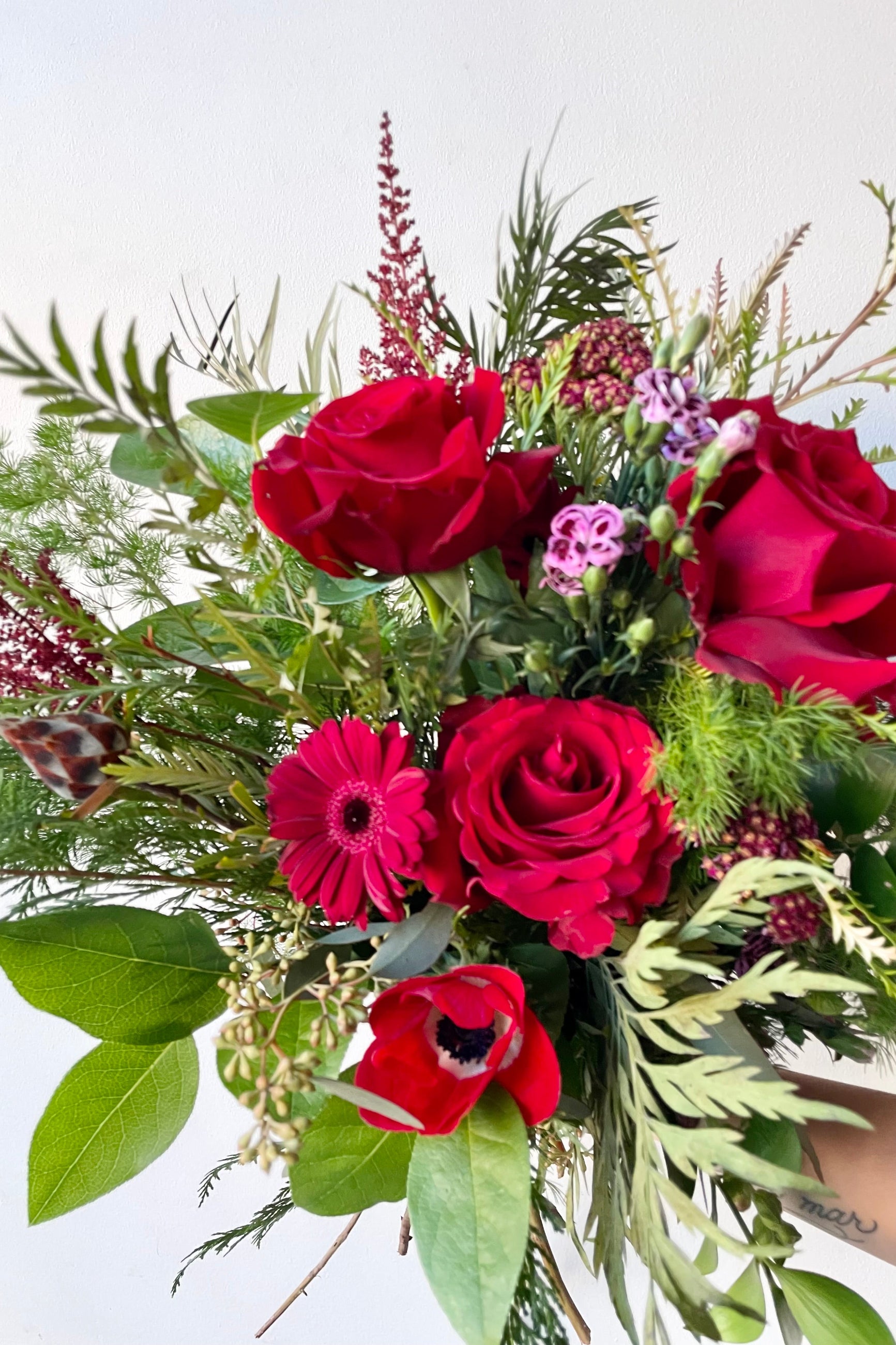 A hand holds an example of fresh Floral Arrangement Solstice with ferns and red roses from Sprout Home Floral in Chicago #size_$130