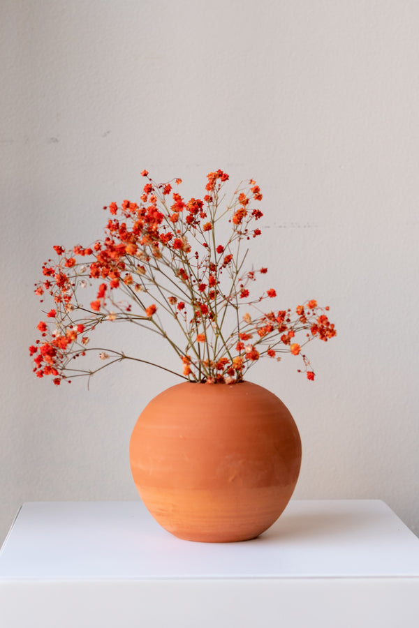 One small round terra cotta vase sits on a white surface in a white room. There are stems of small orange flowers in the vase. It is photographed straight on.
