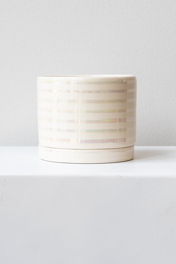 A medium white ceramic planter sits on a white surface in a white room. The planter has thin stripes of iridescent glaze and a matching drainage tray. The planter is empty. It is photographed straight on.