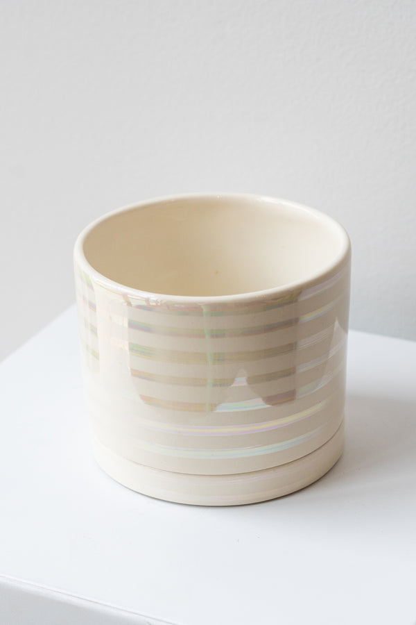 A medium white ceramic planter sits on a white surface in a white room. The planter has thin stripes of iridescent glaze and a matching drainage tray. The planter is empty. It is photographed closer and at an angle.