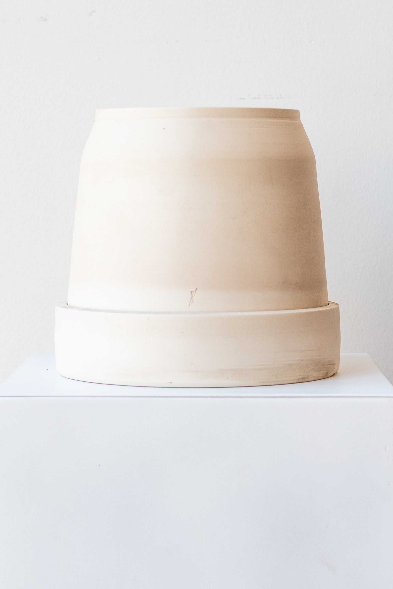 One cream colored stoneware planter sits on a white surface in a white room. The planter is round and squat, with a small logo imprinted in the clay on the bottom. The planter also sits on a round drainage tray. It is photographed straight on.
