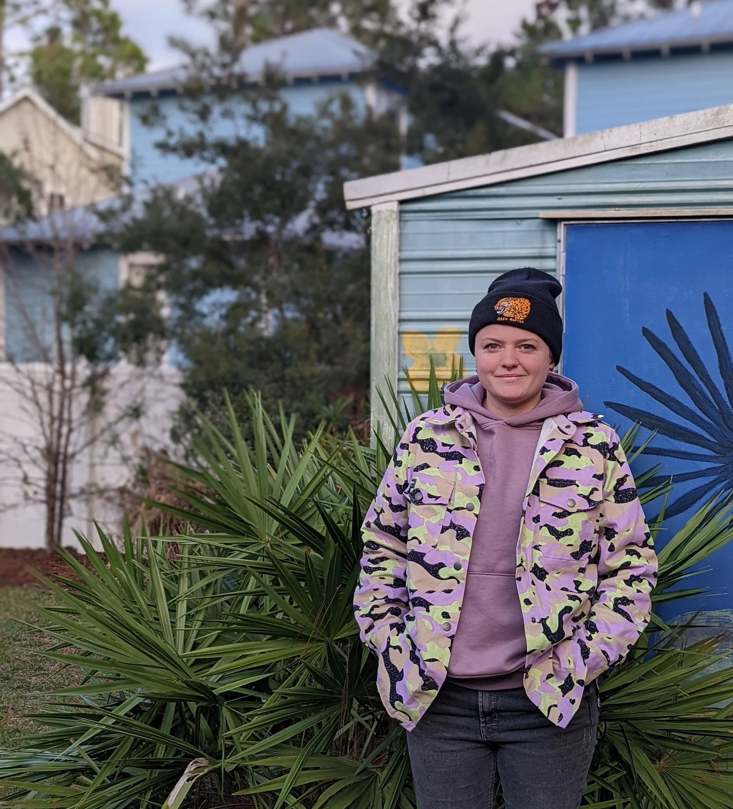 The owner of City in Bloom Ceramics standing in front of a blue garage and plants with a pink army cost and hat ©City of Bloom Ceramics