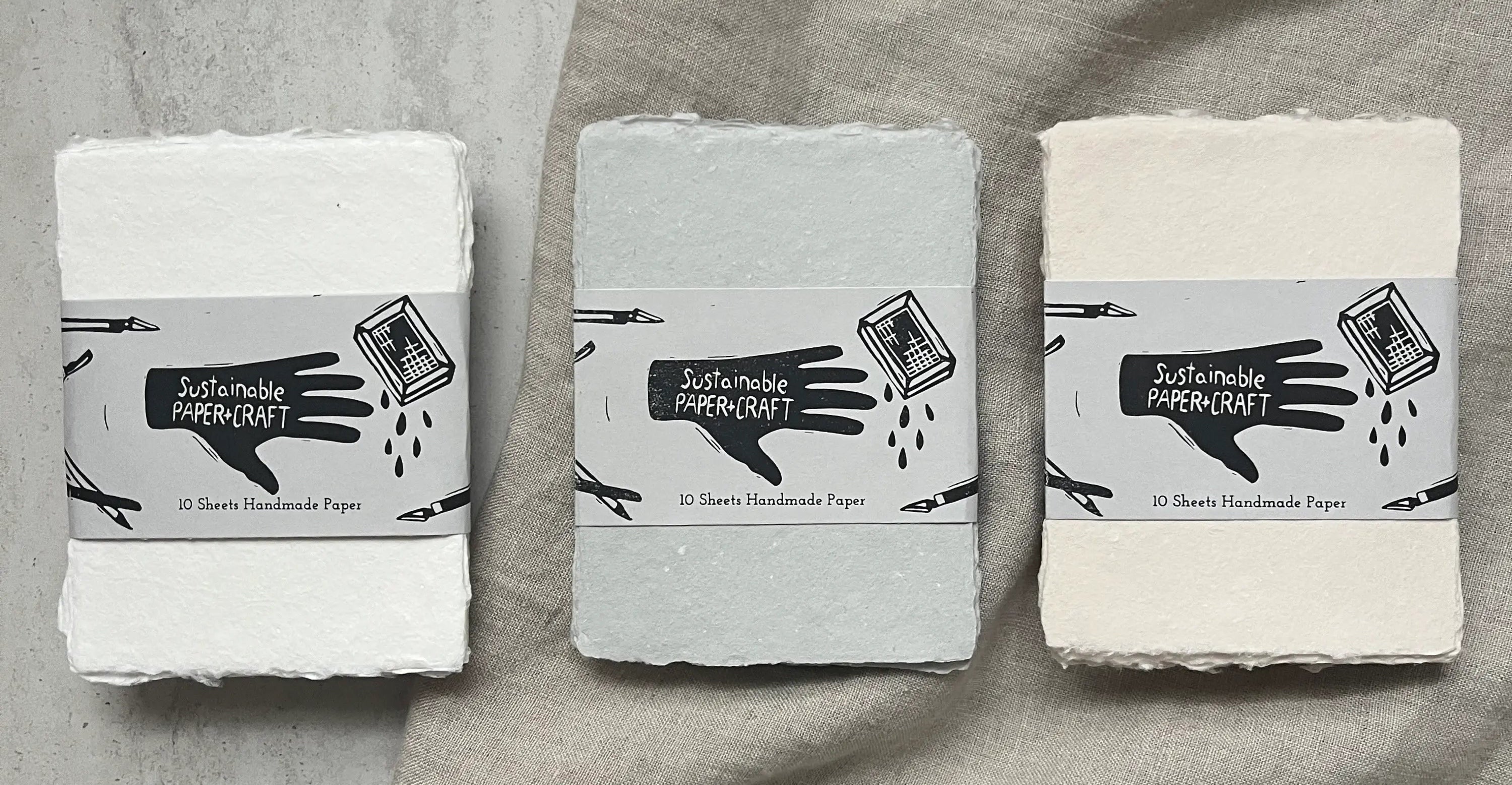 Three rectangular packs of sheet handmade paper in white, light gray, and off-white, each wrapped with a gray paper band labeled 'Sustainable Paper Craft' and '30 Sheets Handmade Paper,' arranged on a draped linen fabric background. ©Sustainable Paper+Craft