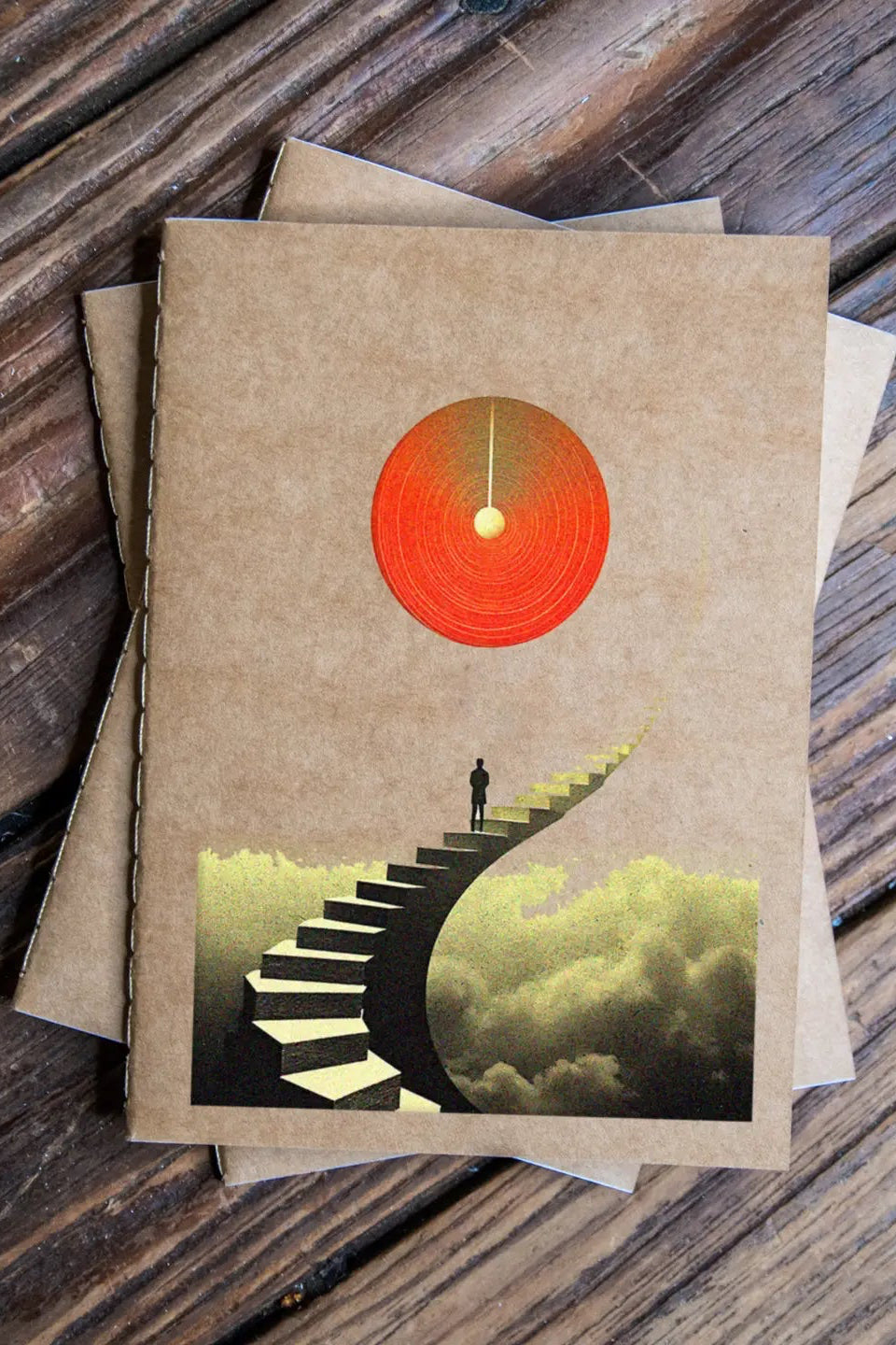 This is Surreal Pack Travel Journal 3 Pack Notebooks with a design of a staircase leading to a red sun on a wooden surface ©Glum Lot Press
