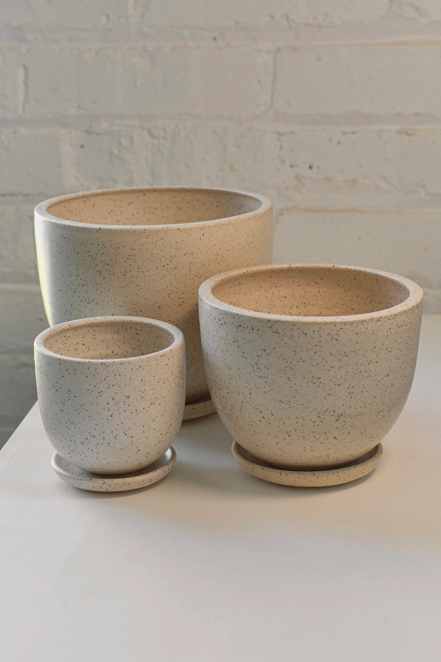 Three ceramic cream speckle tabletop pots of varying sizes on a white surface with a light brick wall background. ©Little Fire Ceramics