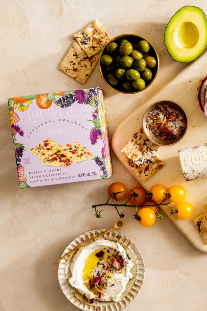 Assorted snacks and appetizers on a wooden board with a box of new Aperitif crackers. ©Flouwer Co.