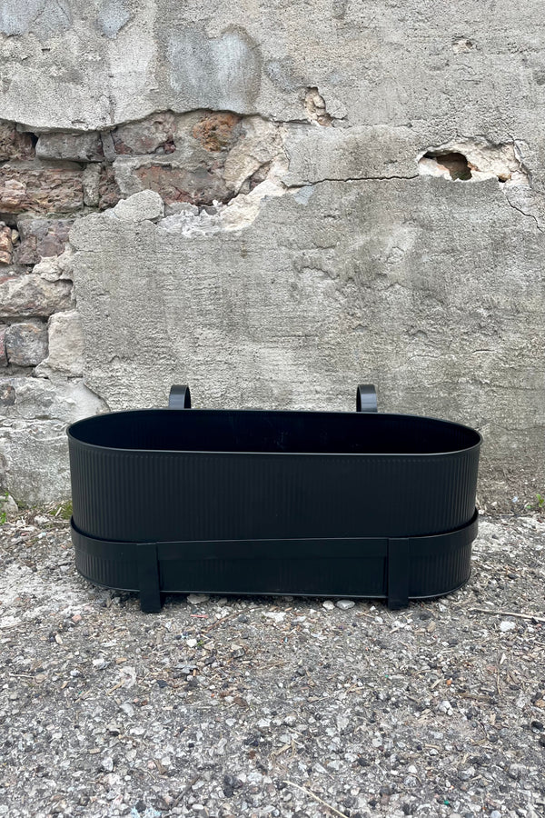 Photo of Ferm Living's Bau Balcony Box in black. The planter features a horizontal holder and bracket and is oval in overall shape. There is a vertical texture to the metal planter. Photo is taken showing the sides and top / inside of the planter. It is photographed on the ground against a cement wall.