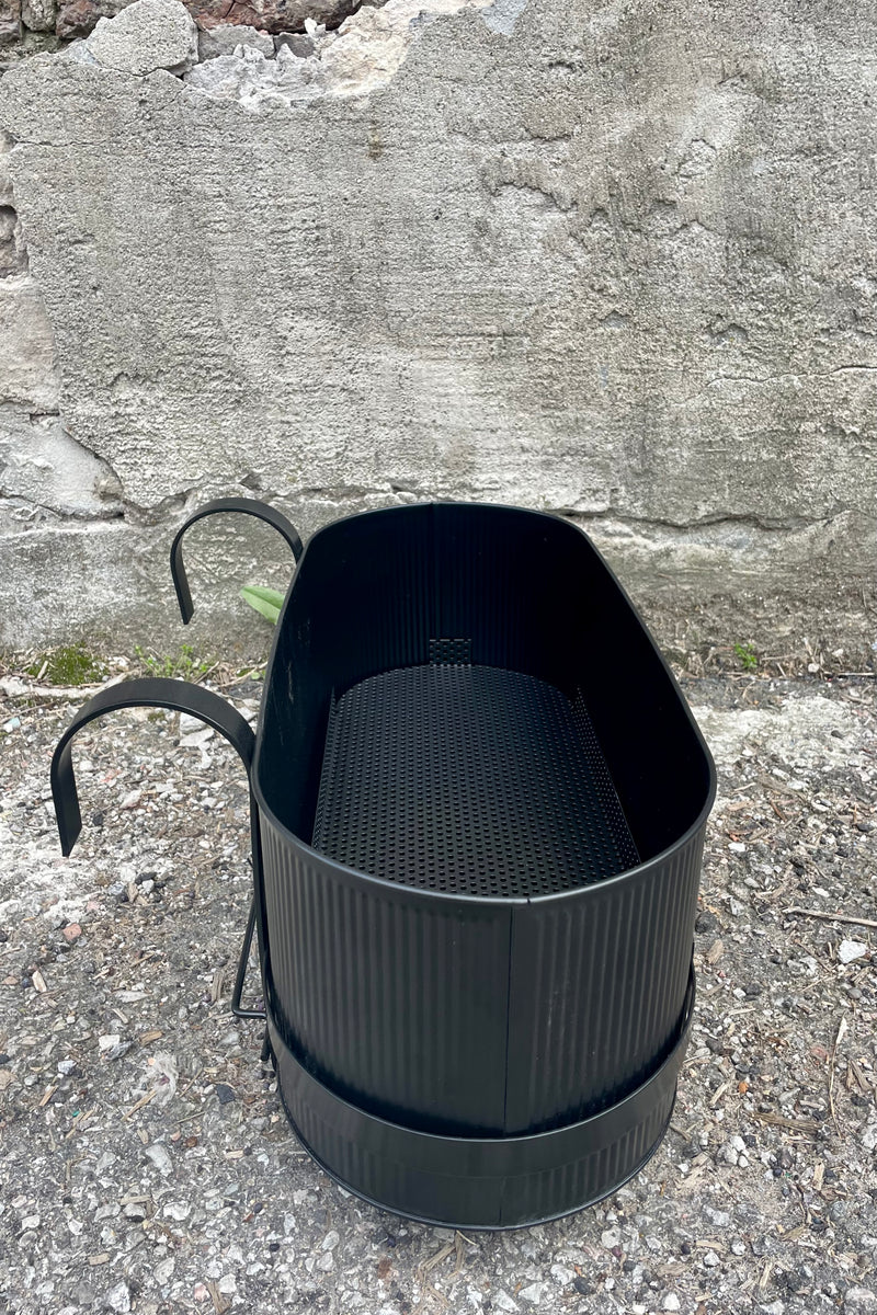 Photo of Ferm Living's Bau Balcony Box in black. The planter features a horizontal holder and bracket and is oval in overall shape. There is a vertical texture to the metal planter. Photo is taken showing the sides and top / inside of the planter. It is photographed on the ground against a cement wall. The view is from the size showing the narrow shape of the planter.