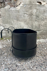 Photo of Ferm Living's Bau Balcony Box in black. The planter features a horizontal holder and bracket and is oval in overall shape. There is a vertical texture to the metal planter. Photo is taken showing the sides and top / inside of the planter. It is photographed on the ground against a cement wall. The view is from the size showing the narrow shape of the planter.