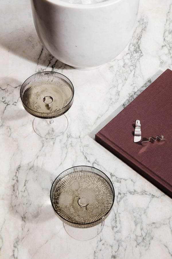 Smoked Champagne saucers by Ferm Living filled with Champagne and shot from above sitting on a marble table with a red book.