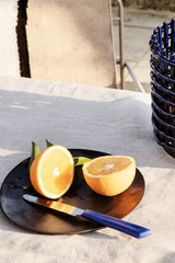 Flow plate in black sitting on a table with a tablcloth and cut orange sitting on the plate with a knife.