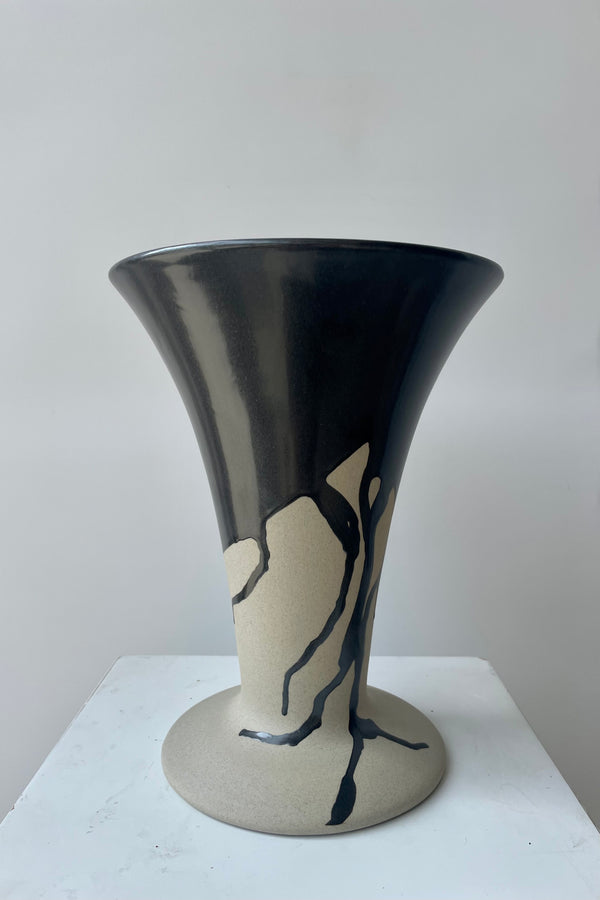 Photo of a Flores vase by Ferm Lliving. The vase is raw beige clay with a black irregular glaze at the rim and extending downward. The vase flares wides at the top and again at the base. The vase is photographed in a white room against a white wall. 