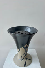 Photo of a Flores vase by Ferm Lliving. The vase is raw beige clay with a black irregular glaze at the rim and extending downward. The vase flares wides at the top and again at the base. The vase is photographed in a white room against a white wall. Shown inside the opening is a ceramic floral frog.
