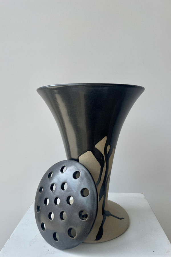Photo of a Flores vase by Ferm Lliving. The vase is raw beige clay with a black irregular glaze at the rim and extending downward. The vase flares wides at the top and again at the base. The vase is photographed in a white room against a white wall. Placed in front of the vase is a black ceramic floral frog.